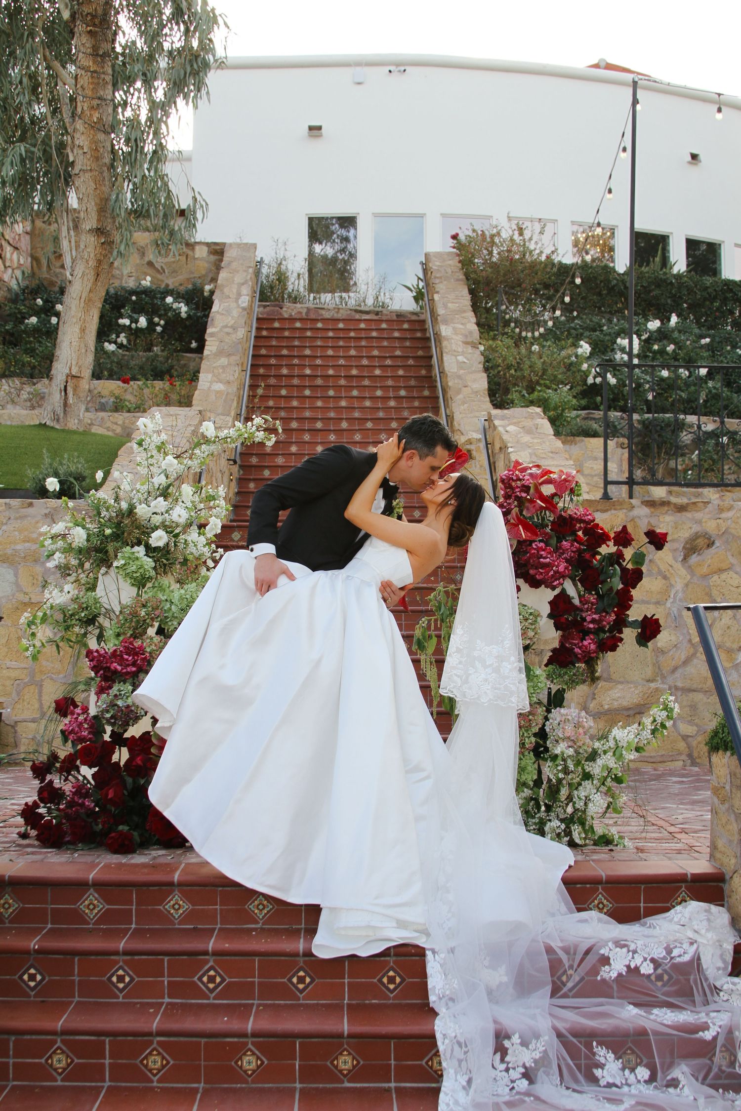 Bride and groom share a romantic dip kiss on flower-adorned brick stairs at an outdoor wedding venue.