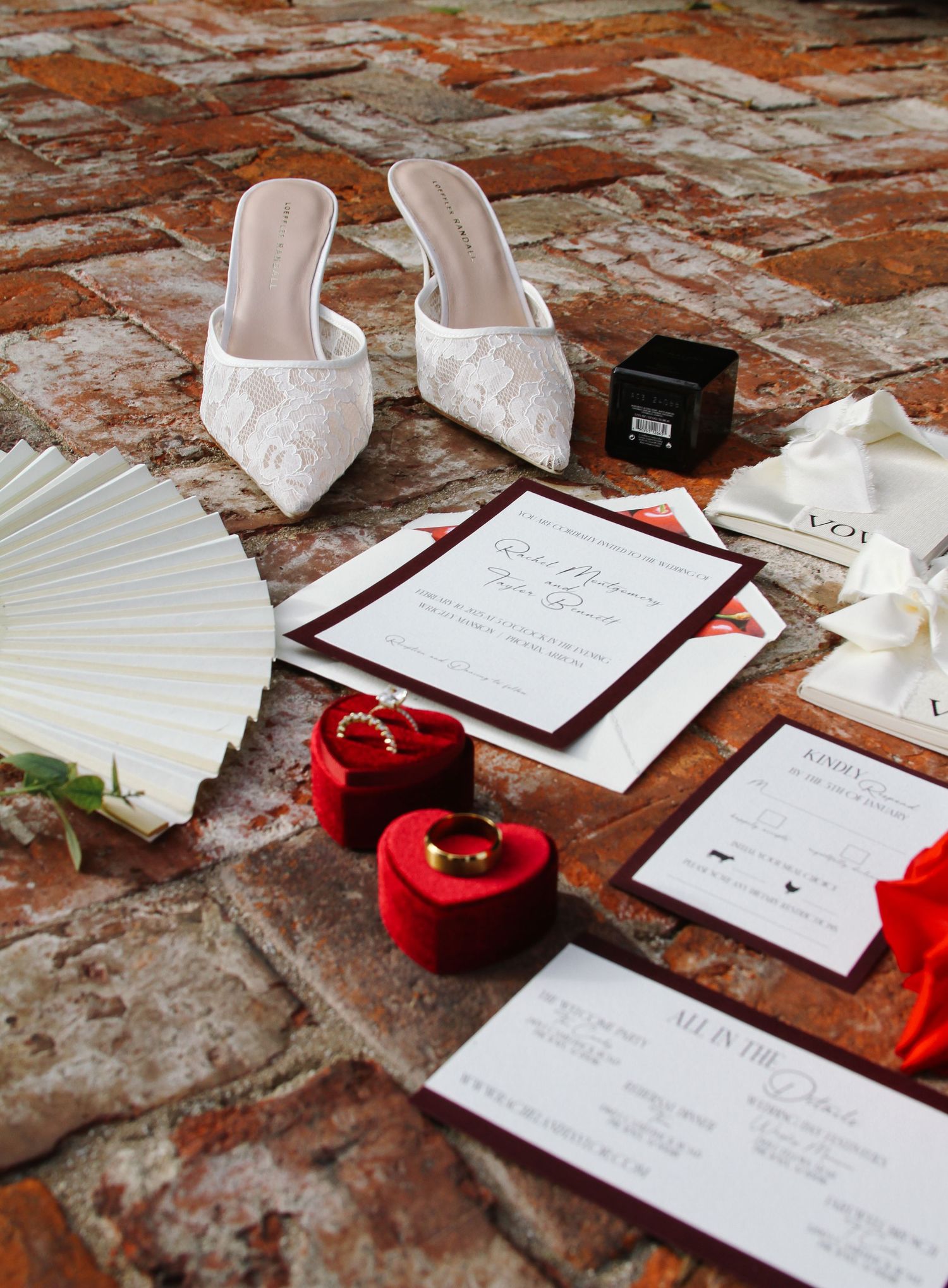 Wedding flat lay with lace heels, red velvet ring boxes, gold bands, and stationery on brick floor.