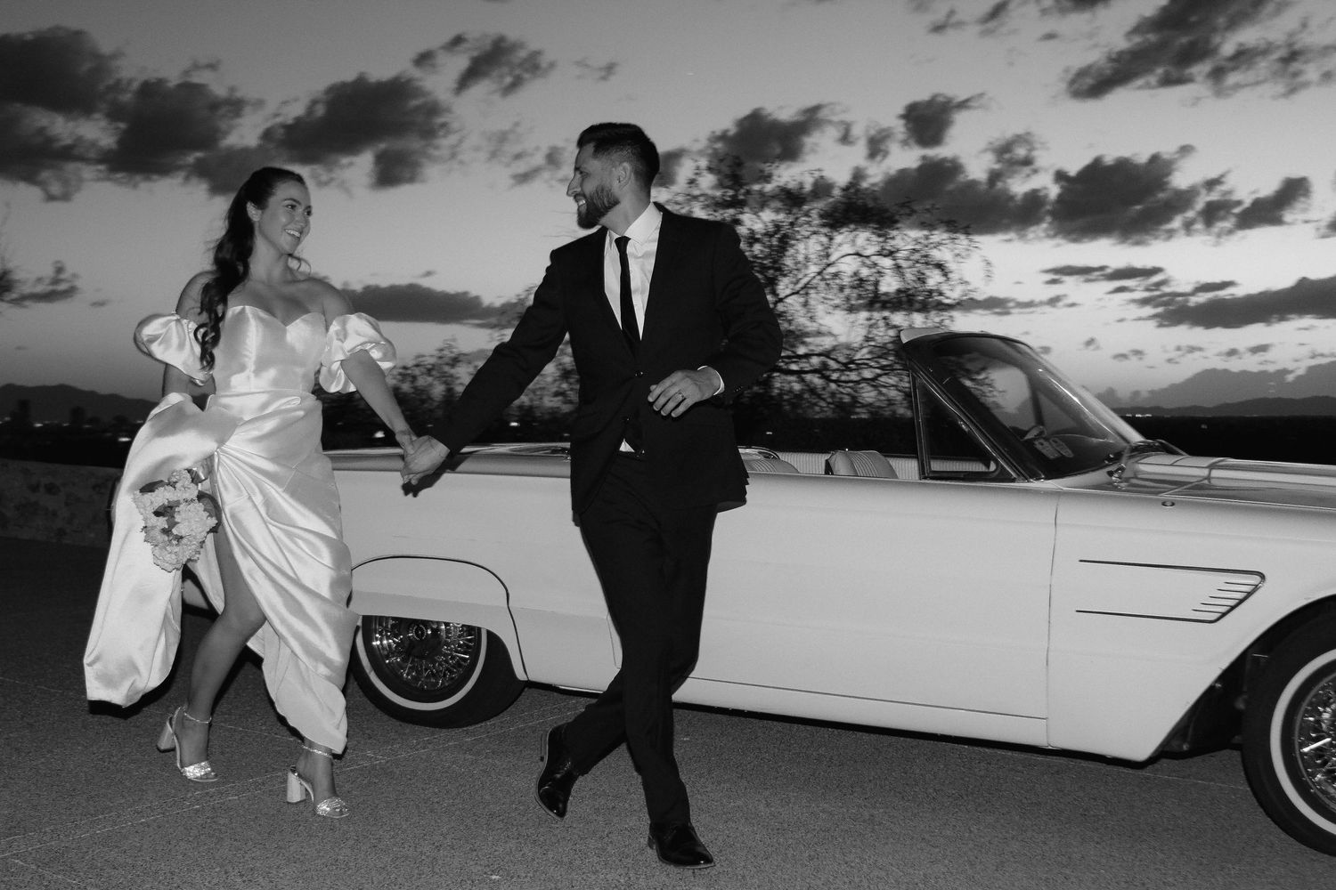 A couple in wedding attire poses by a classic white car at sunset.