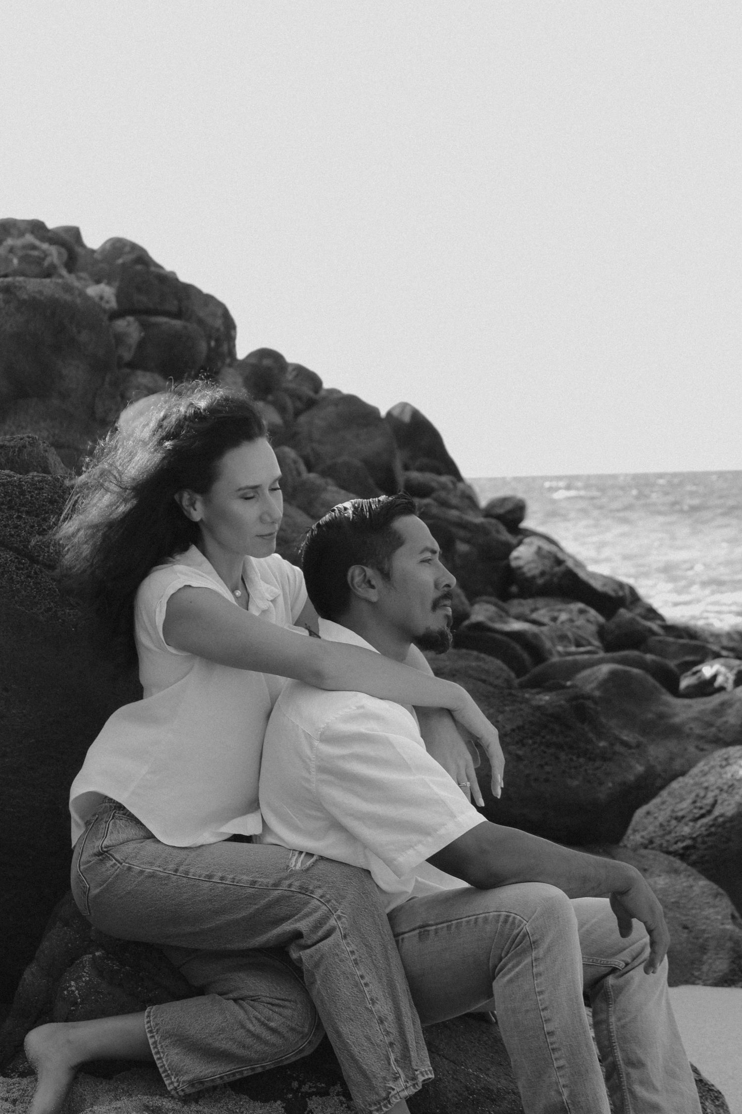 A couple sits close together on coastal rocks by the ocean in a series of black and white romantic images.