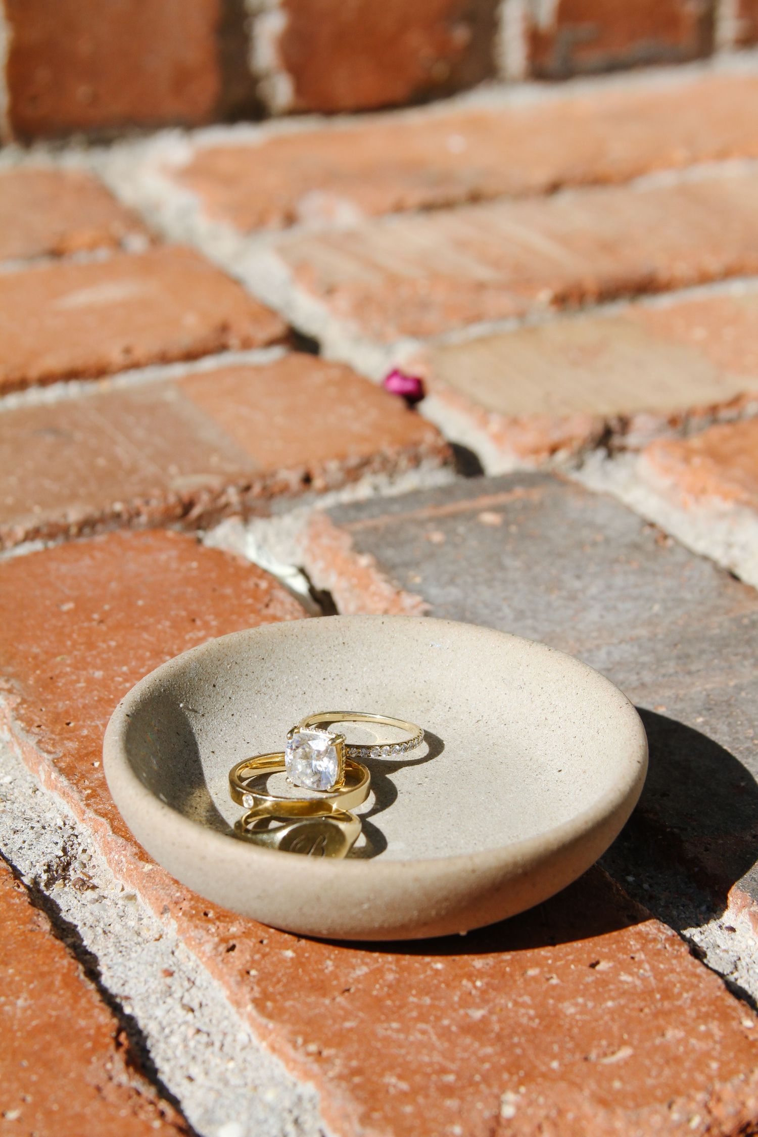 Two gold engagement rings rest in a small beige ceramic dish placed on red brick pavement in natural sunlight.