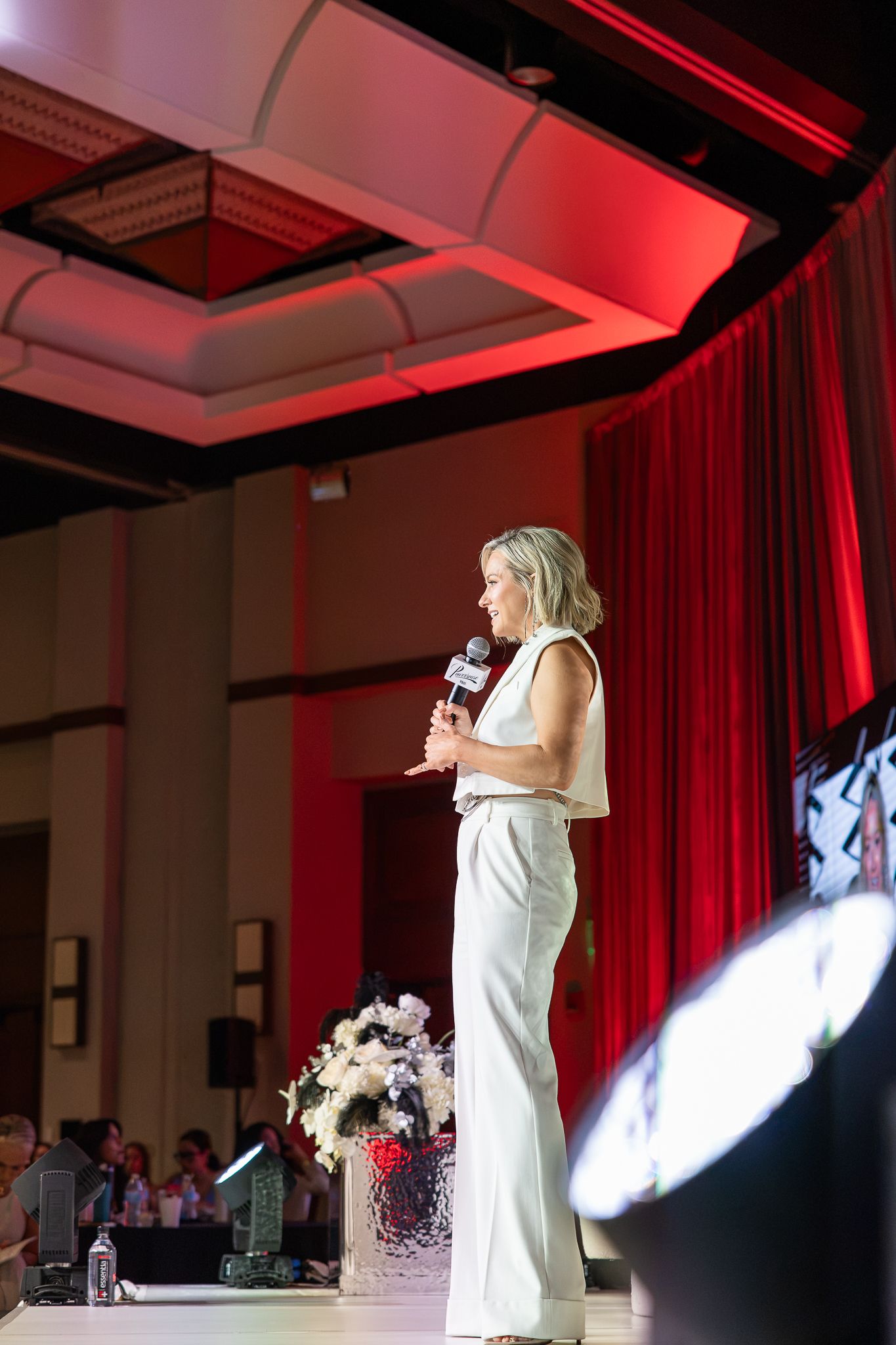 Woman in white outfit speaks on stage with microphone at elegant event with red curtains and dramatic lighting.