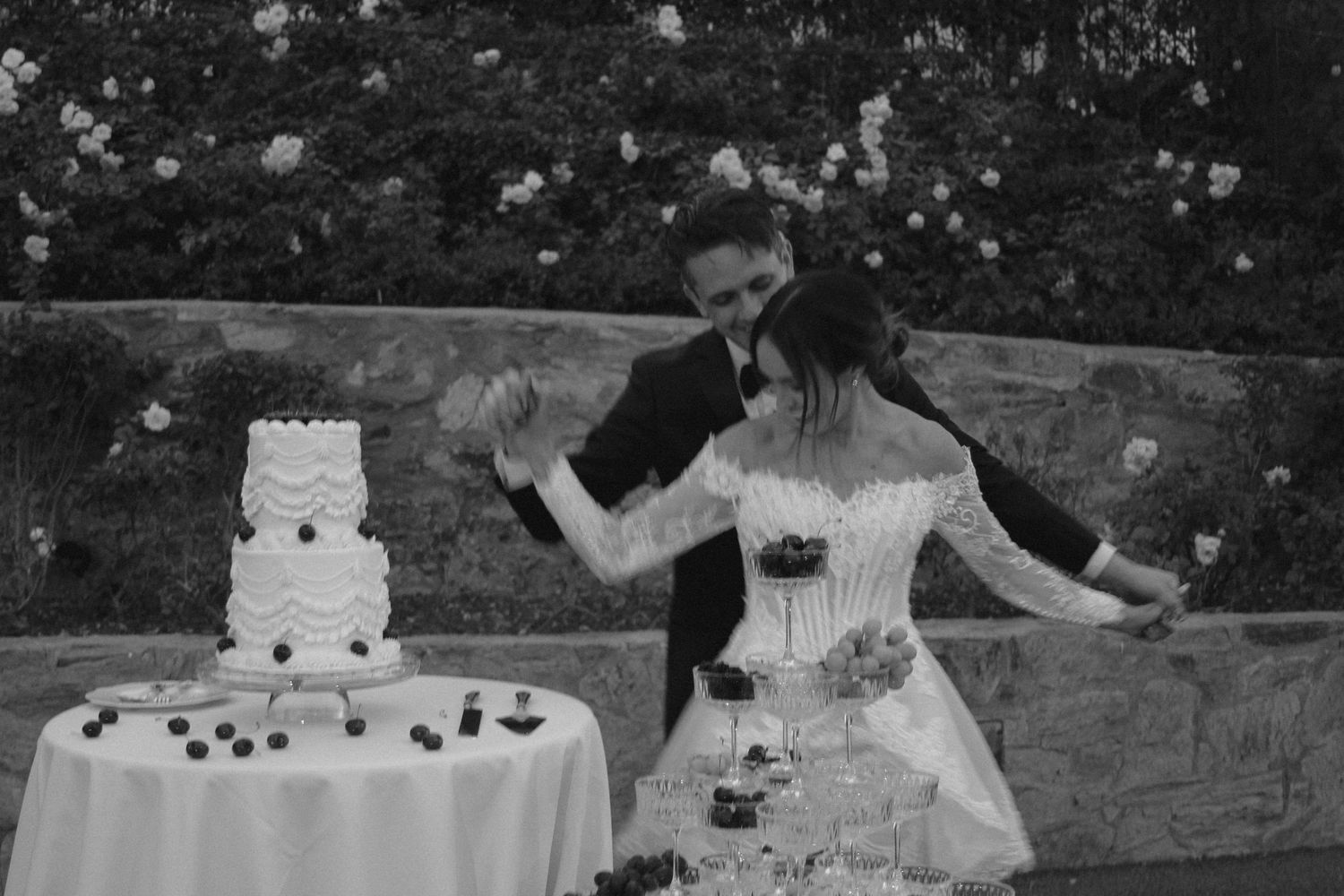 Black and white photo of a bride and groom cutting their tiered wedding cake outdoors near a stone wall.