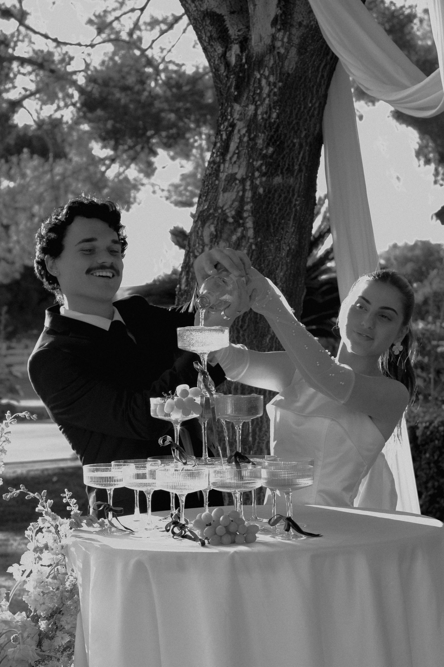 Bride and groom smiling while pouring champagne tower at outdoor wedding reception in black and white photo.