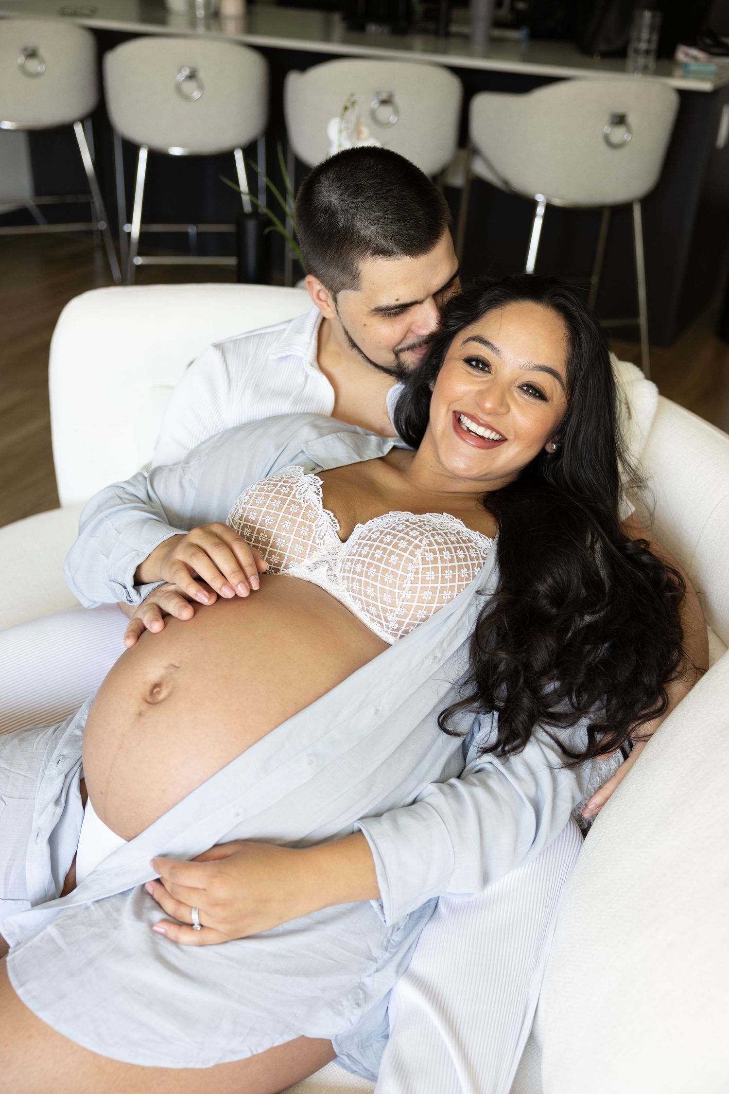 Happy pregnant woman reclines against her partner on a white sofa, showing her baby bump in a cozy home setting.
