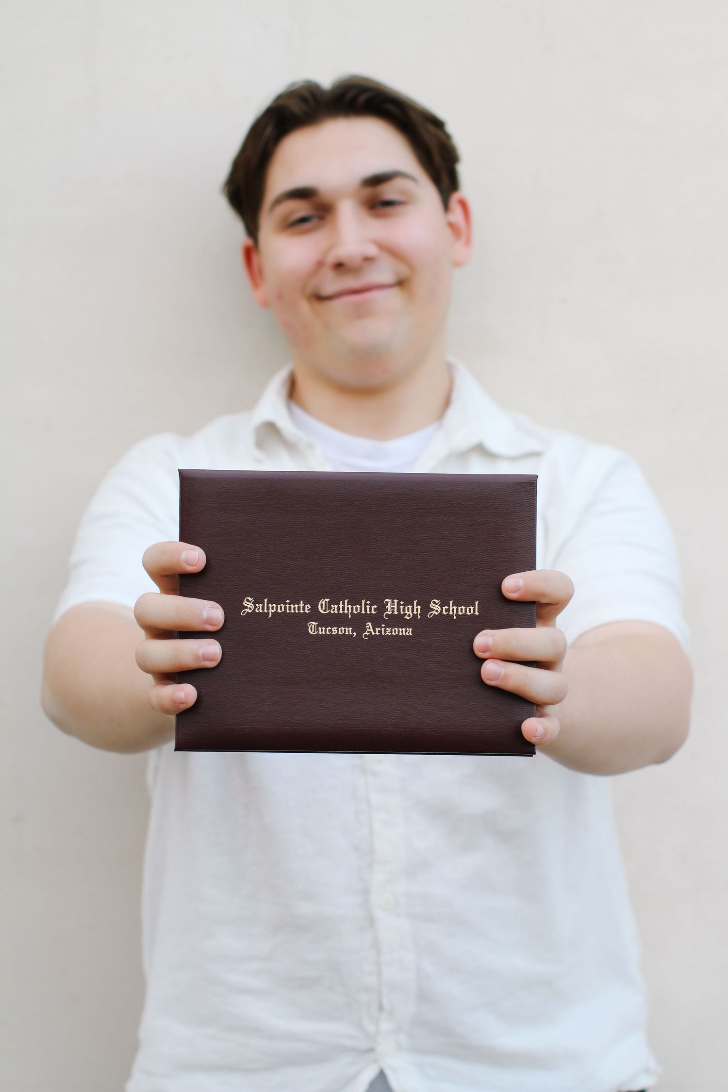 Graduate holding up a dark brown diploma from Saguaro Catholic High School against a light wall.