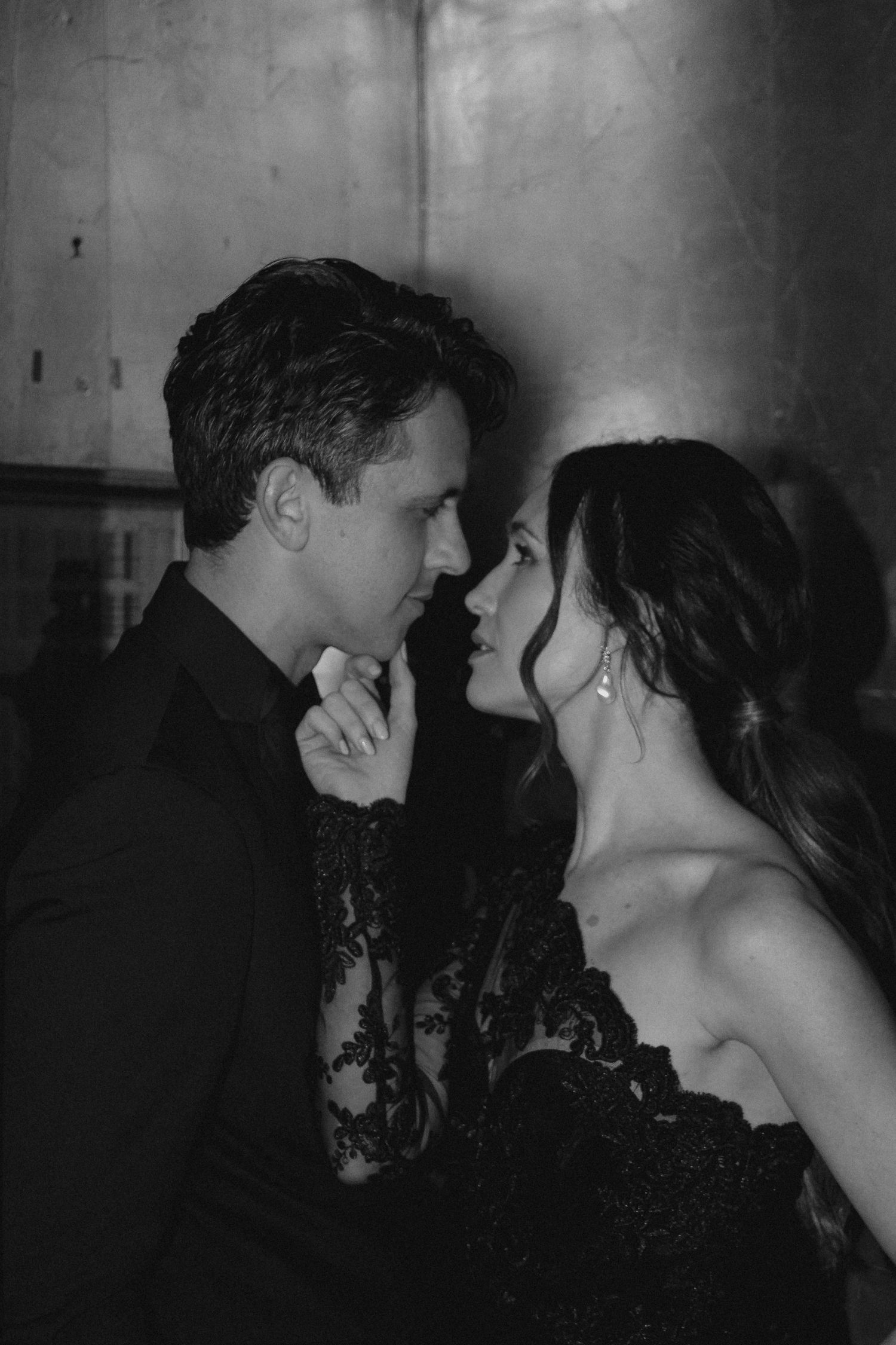 Black and white image of couple in evening wear sharing tender moment against industrial backdrop.