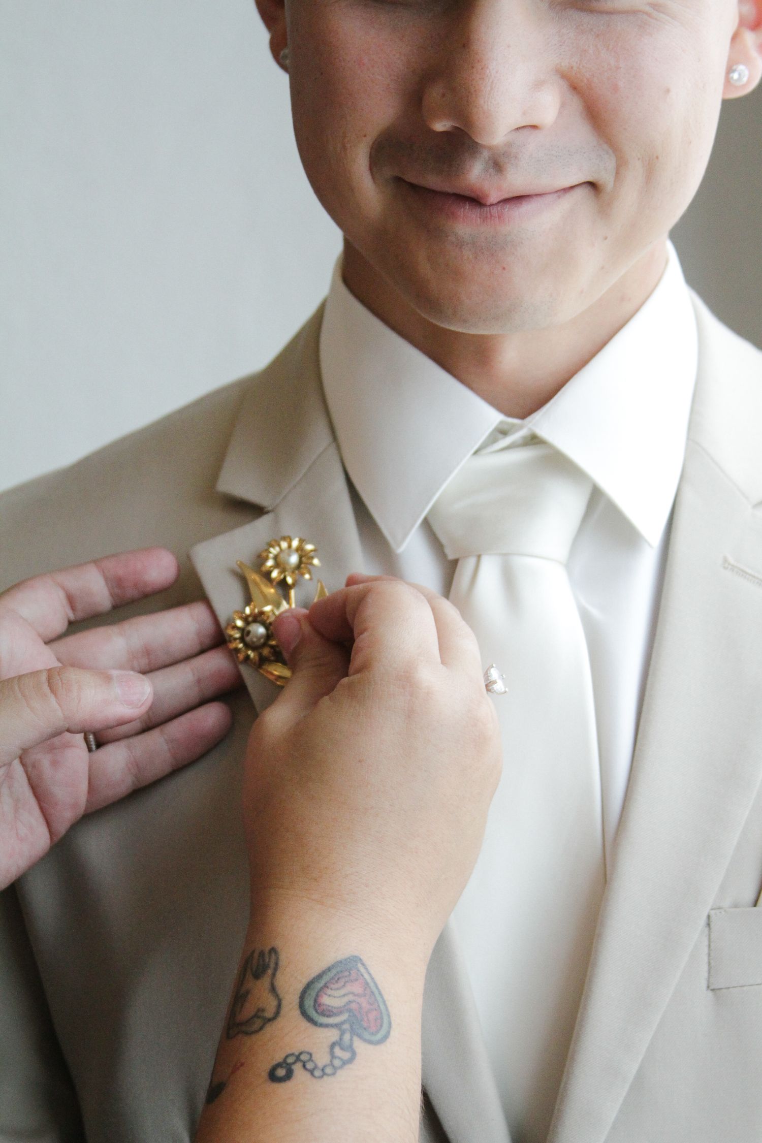 Tattooed hand pins a gold floral boutonniere on a smiling groom in a white suit.