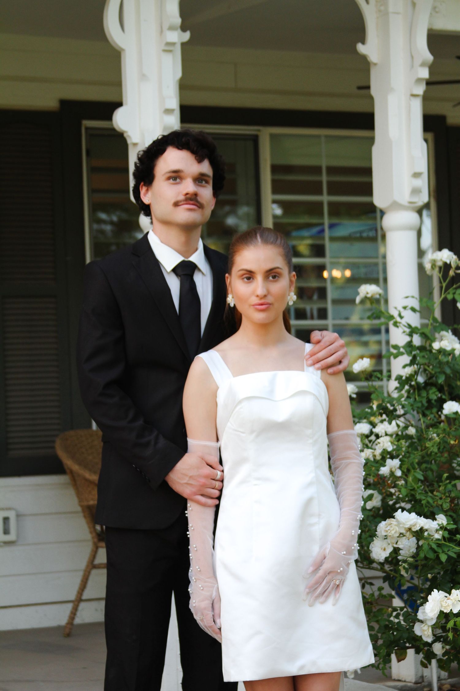 Elegant couple in wedding attire posing on a white porch, man in black suit, woman in white mini dress.