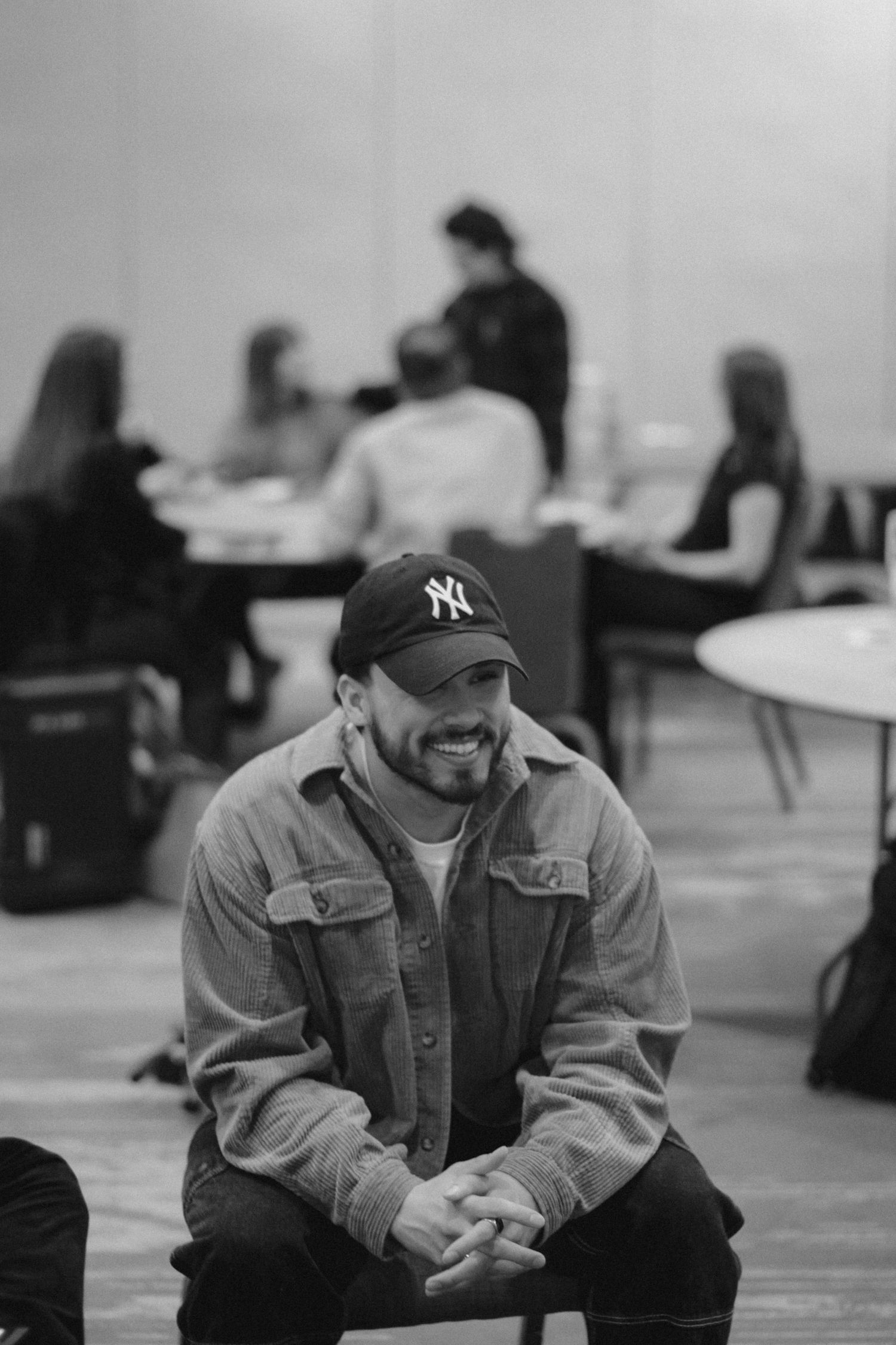 Black and white photo of a smiling man in a NY Yankees cap and denim jacket sitting with hands clasped in a room.