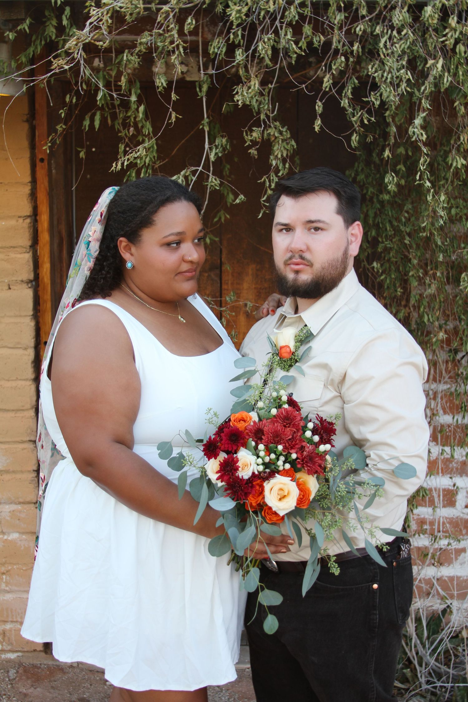 Couple poses with floral bouquet featuring red and peach roses with eucalyptus greenery.