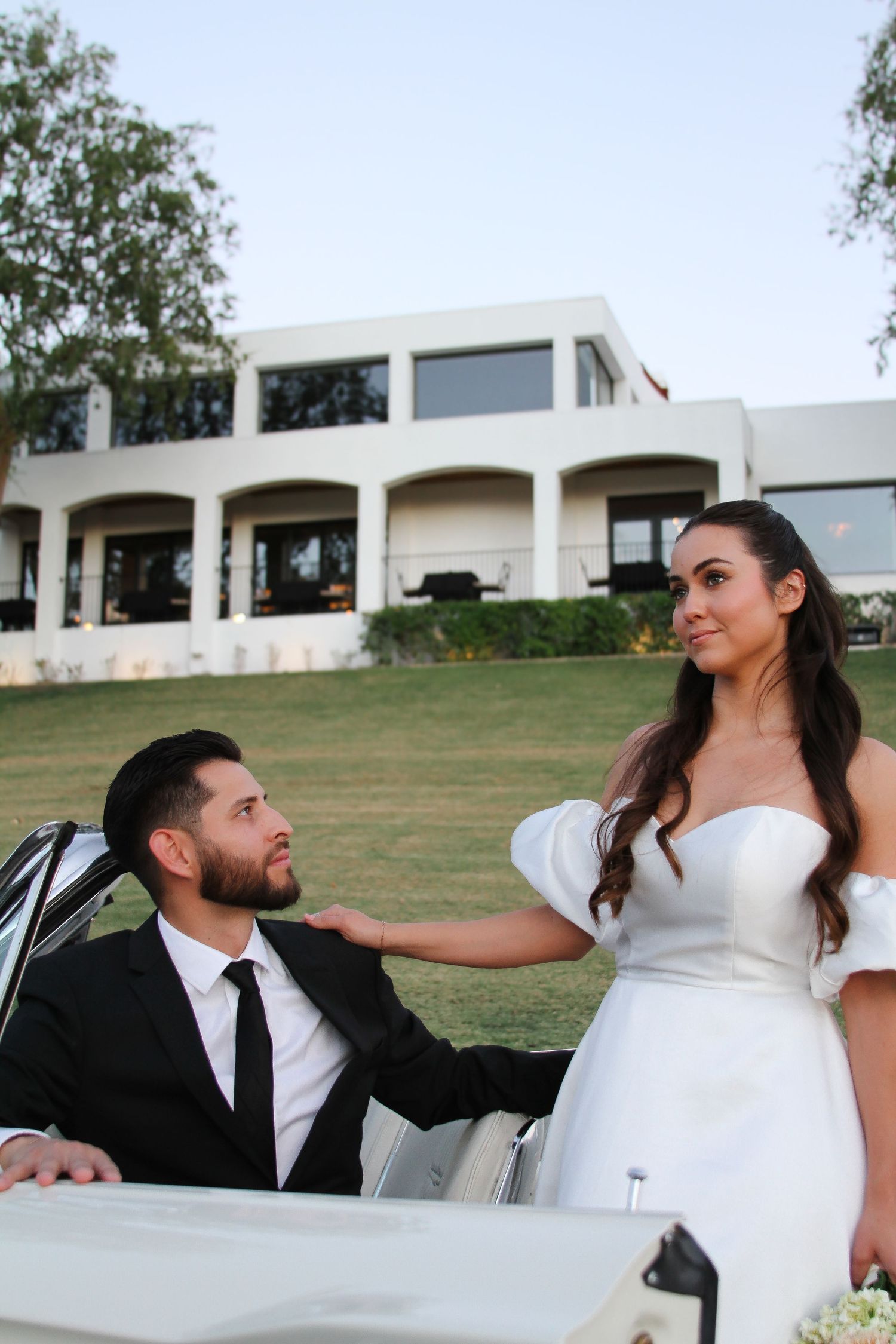 A romantic wedding couple poses beside a vintage car with a white mansion in the background.