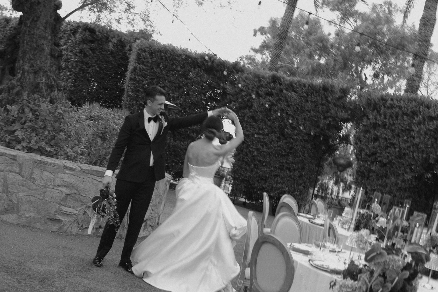 Black and white photo of a bride and groom dancing outdoors at an elegant wedding reception with lush hedges.