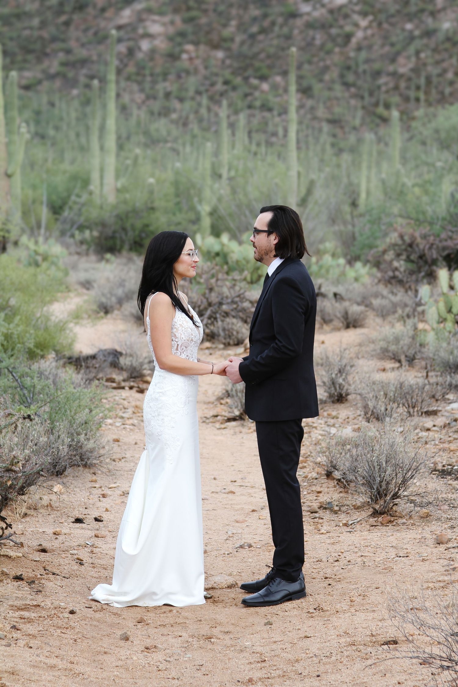 A couple in wedding attire shares a moment on a desert trail surrounded by towering saguaro cacti.