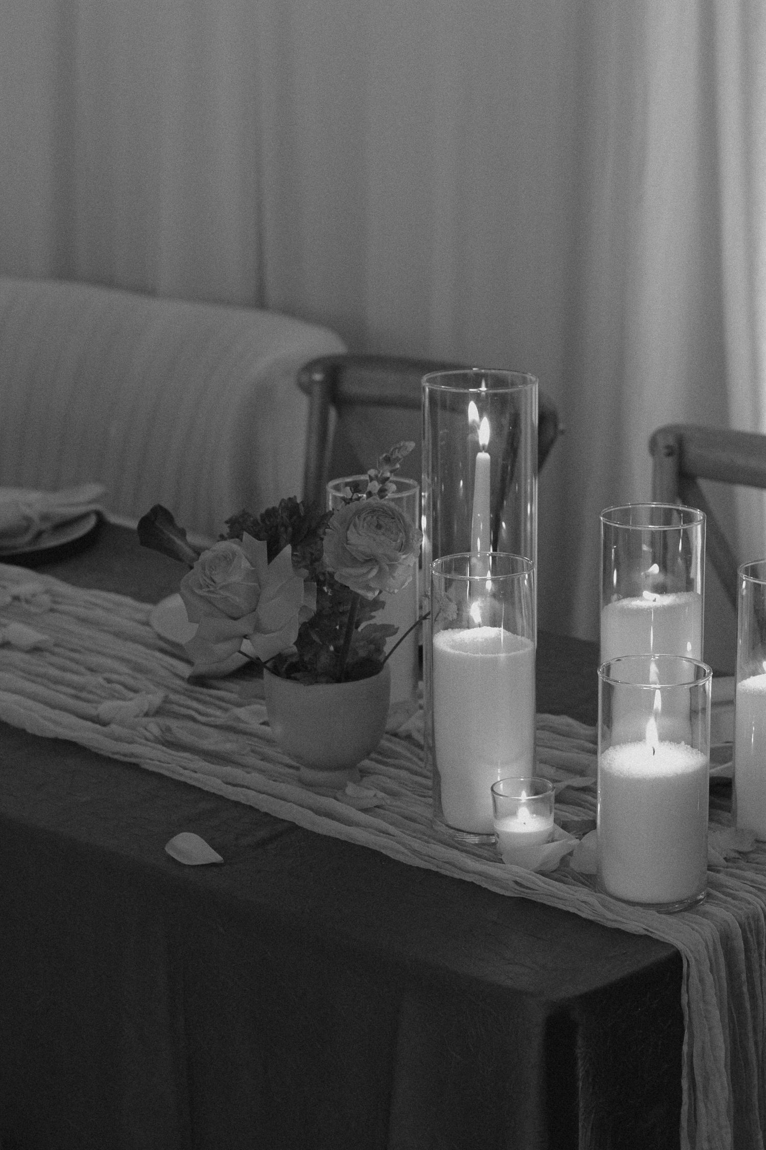 Tyler Garlej Photography - A series of lit white candles in glass holders arranged on a wooden surface with fabric in a monochrome setting.