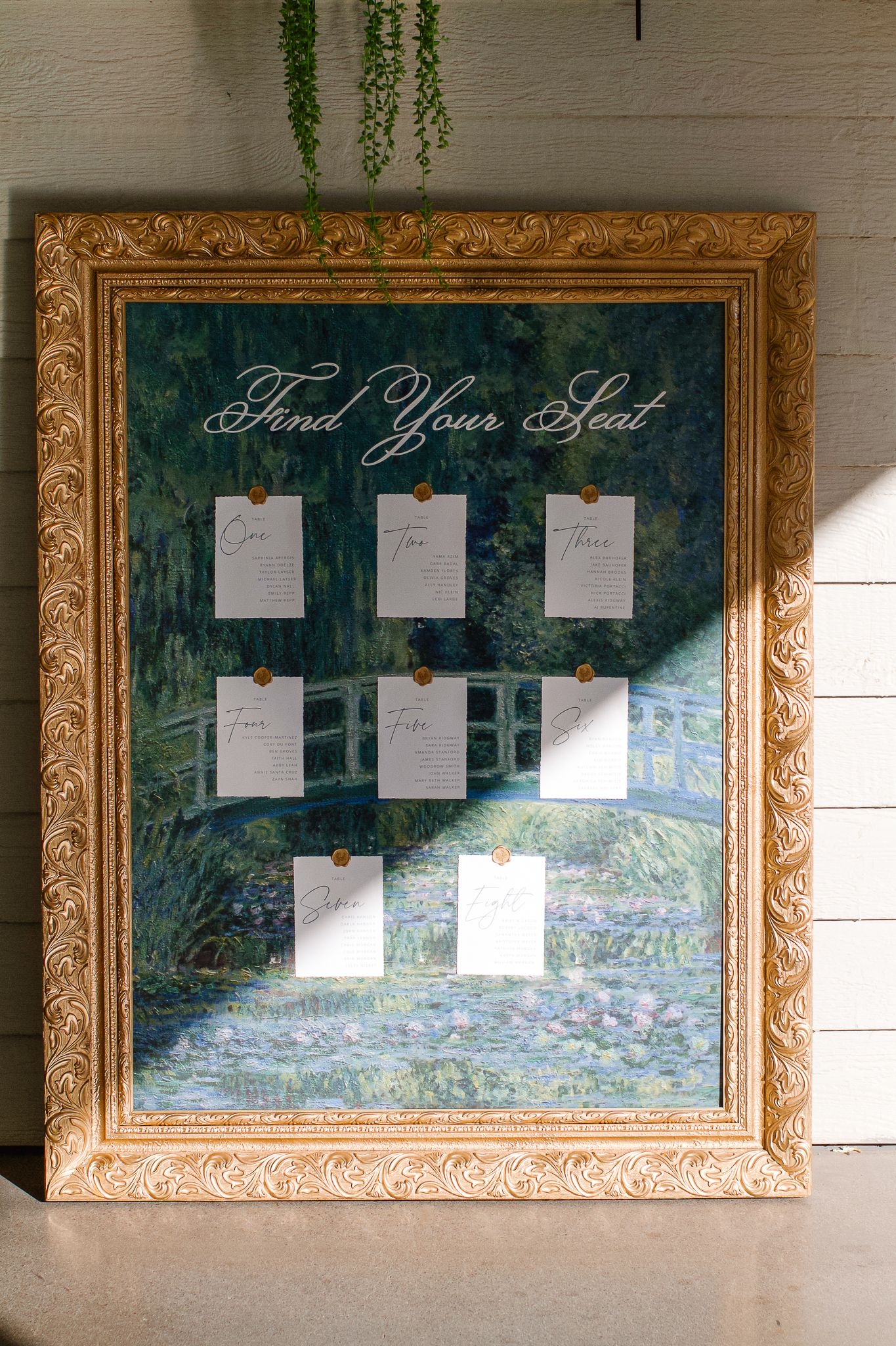 A wedding seating chart displayed across three ornate gold frames with white papers arranged on a painted landscape backdrop.