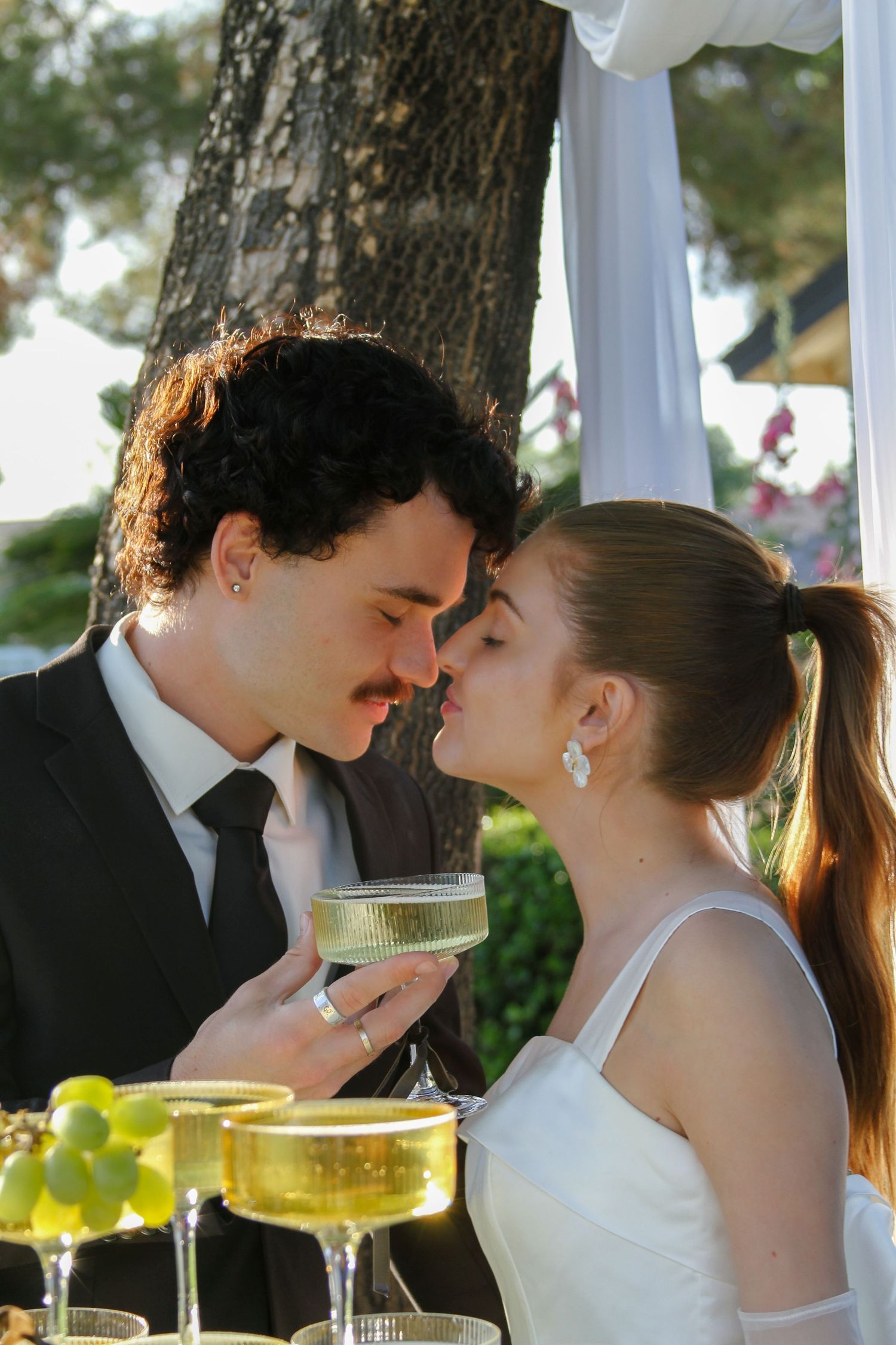 Newlywed couple sharing a romantic kiss at outdoor wedding reception, holding champagne coupe glasses.