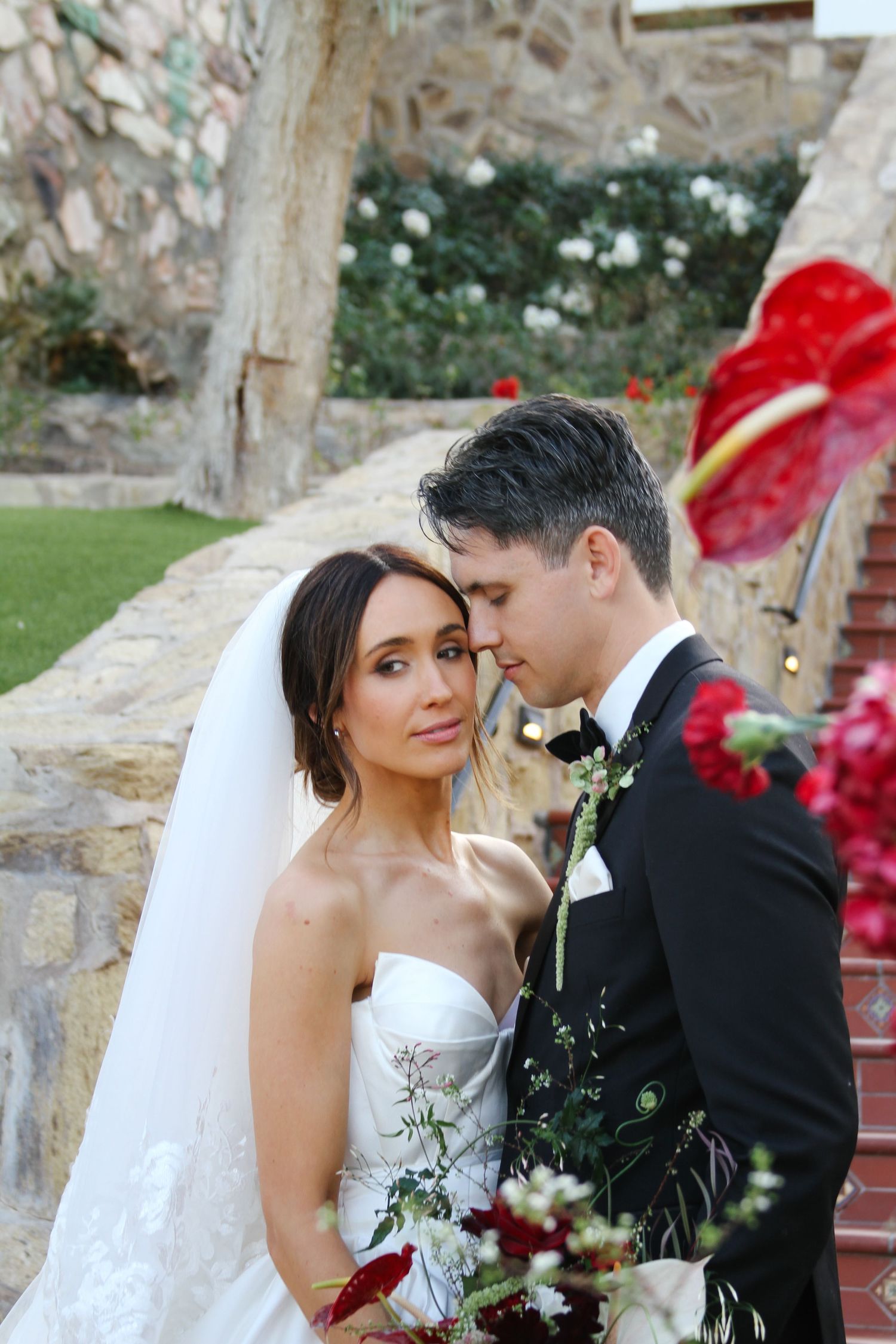 Bride in strapless white gown with veil holds red bouquet as groom in black tuxedo leans close outdoors.