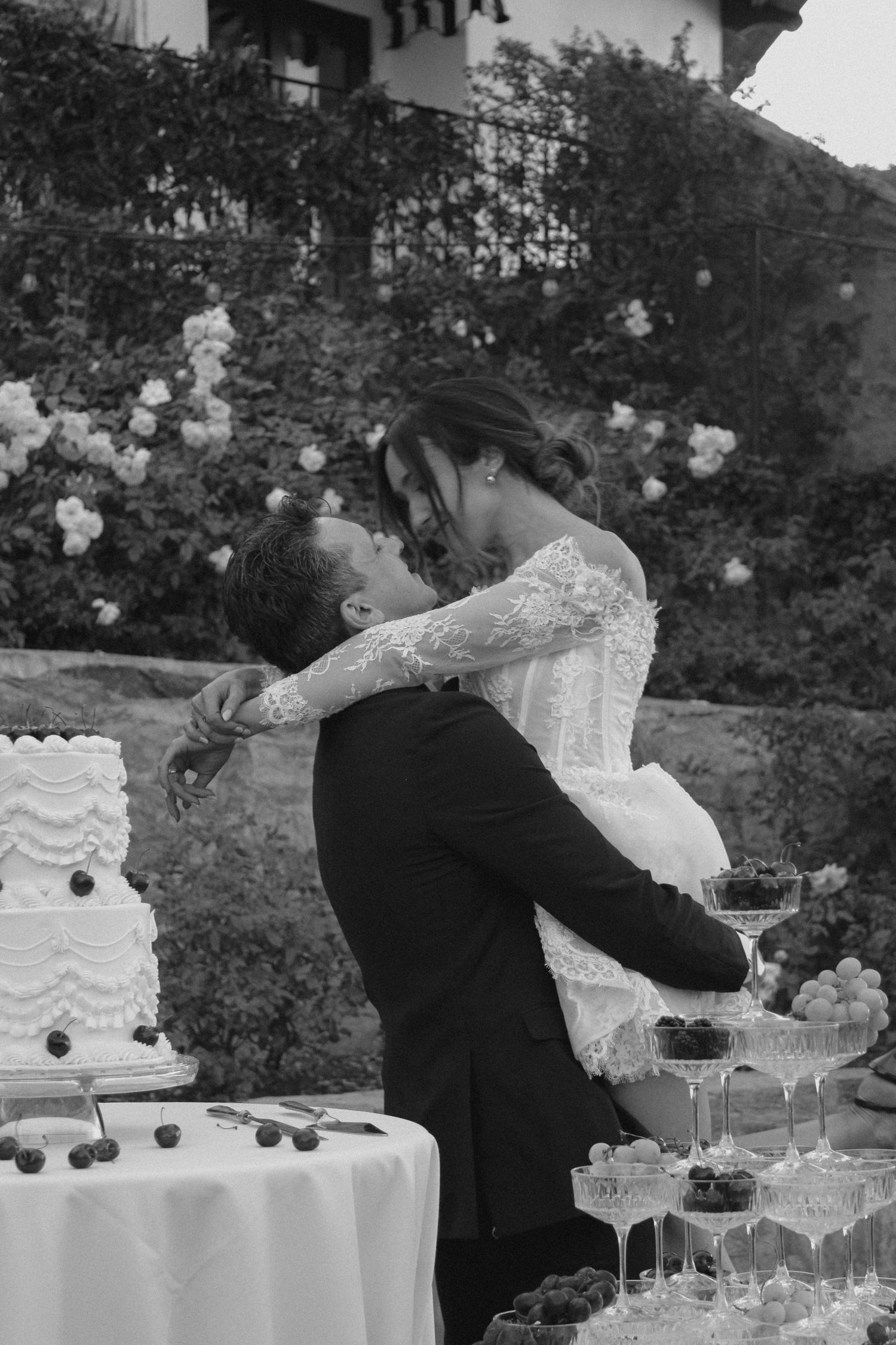 Black and white photo of bride and groom sharing a kiss beside their wedding cake and champagne tower.