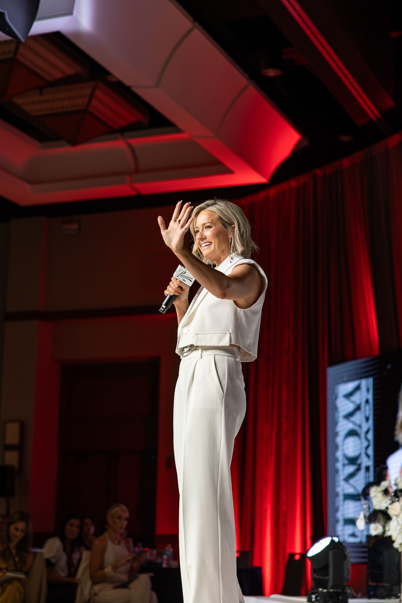 Woman in white jumpsuit speaking on stage with microphone, waving at audience in red-lit ballroom.