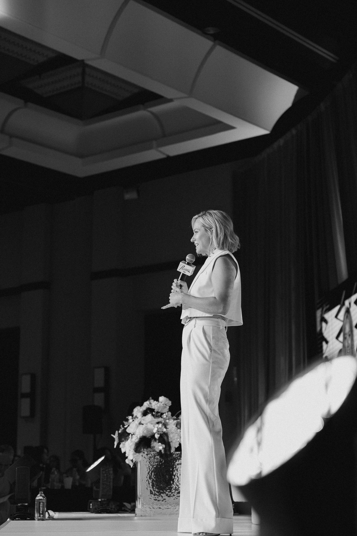 A woman in white outfit speaks into a microphone on stage at an elegant event in black and white photo.