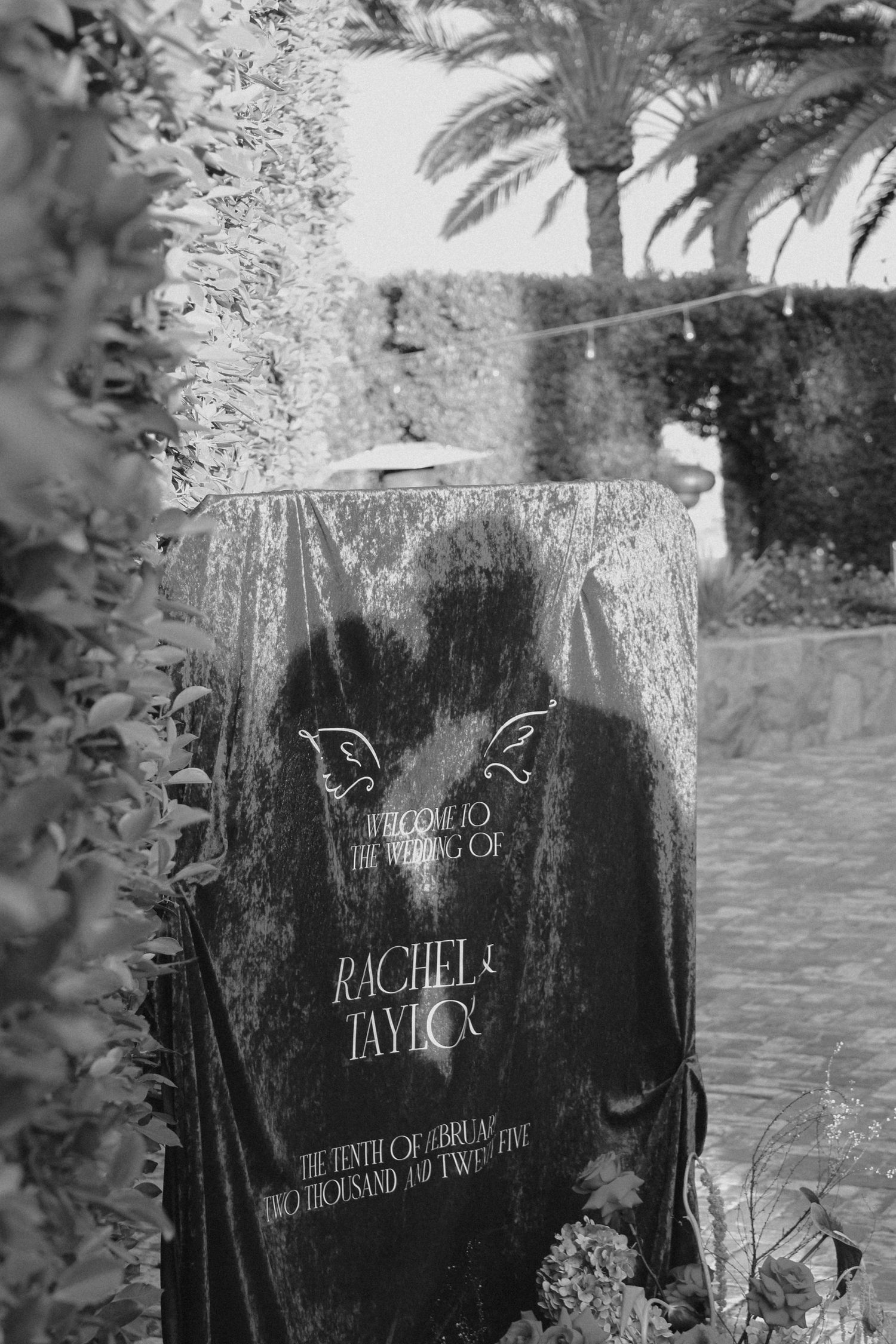 Black and white photo of an elegant wedding welcome sign with palm trees in the background.