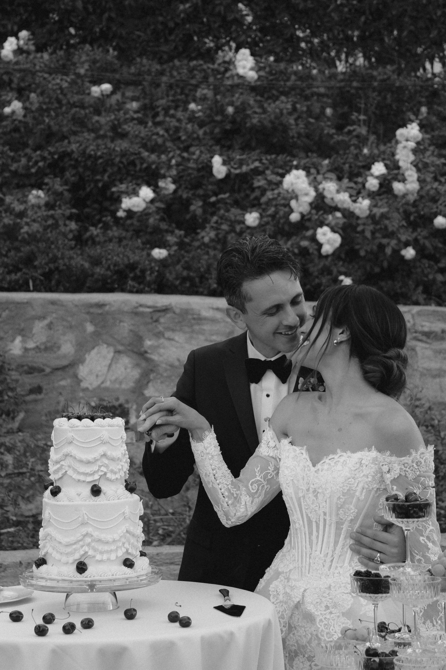 Bride and groom cutting white tiered wedding cake decorated with cherries outdoors.