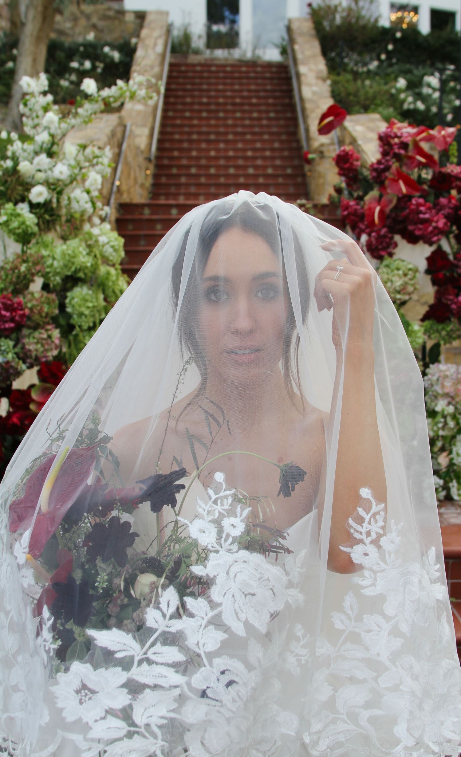 Beautiful bride lifts lace-embroidered veil while holding dark floral bouquet at outdoor garden wedding ceremony.