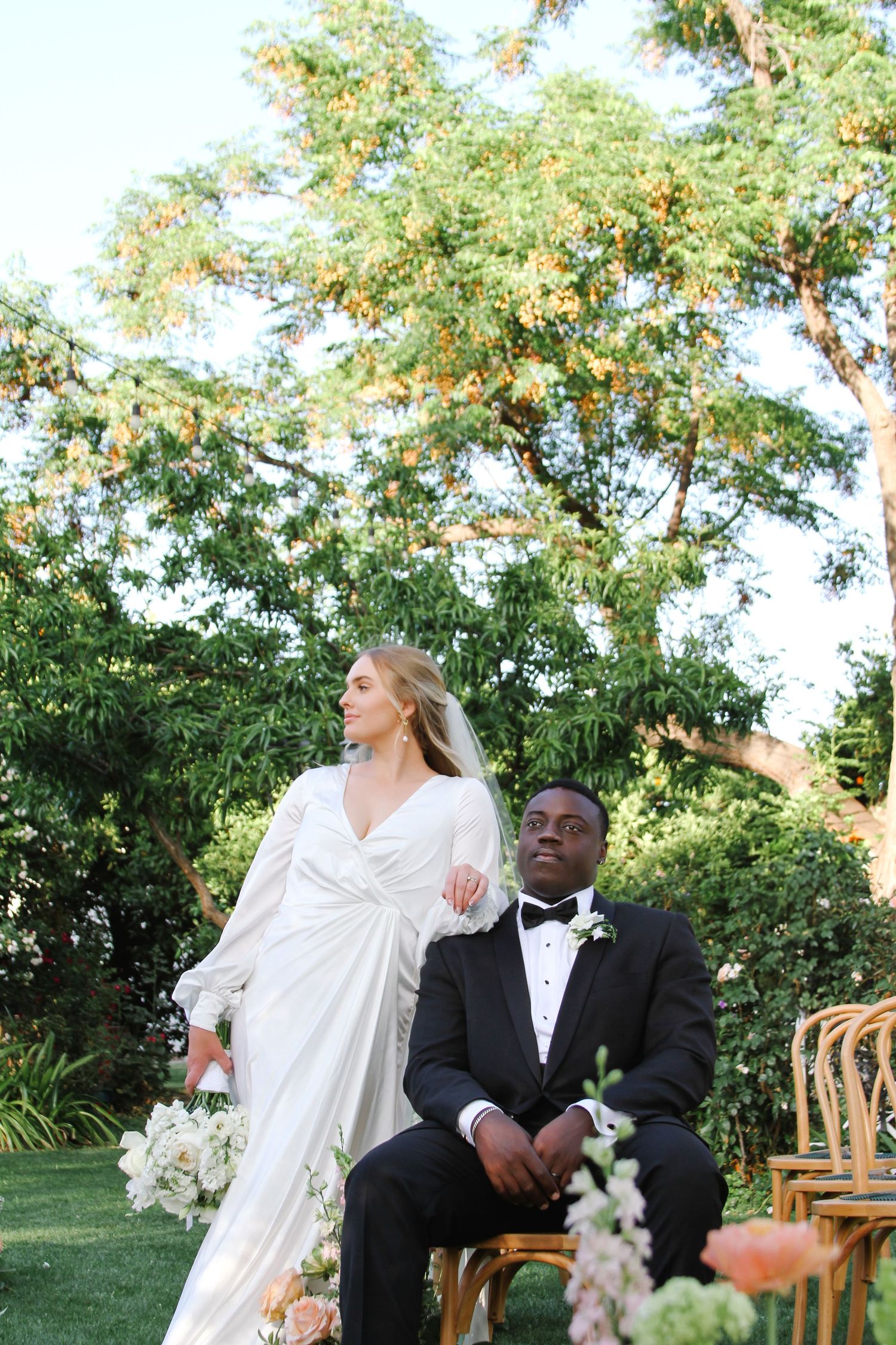 Couple in wedding attire sharing a joyful moment under green trees during an outdoor ceremony.