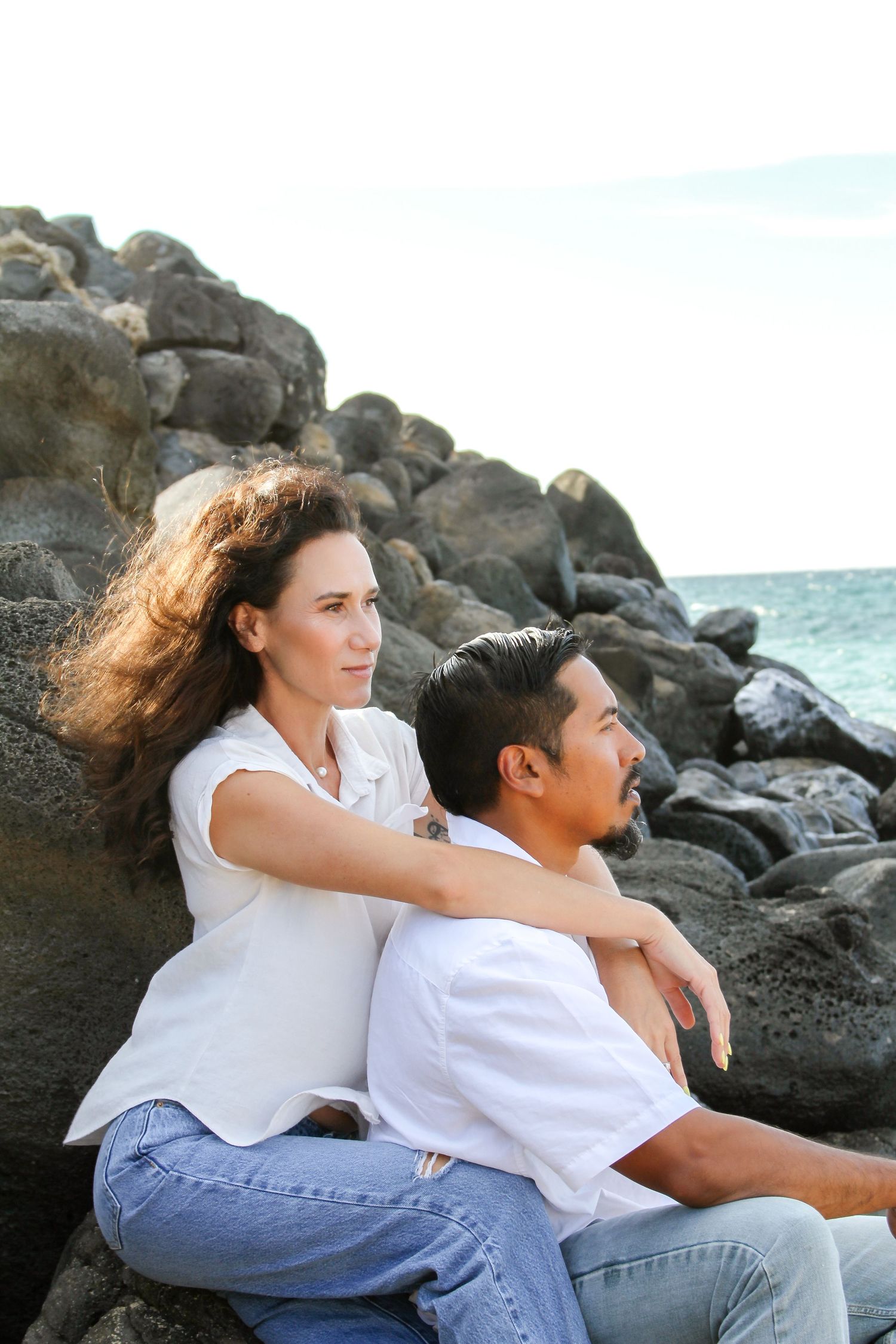 Couple in white shirts sitting together on rocky beach shore as wind blows through their hair.