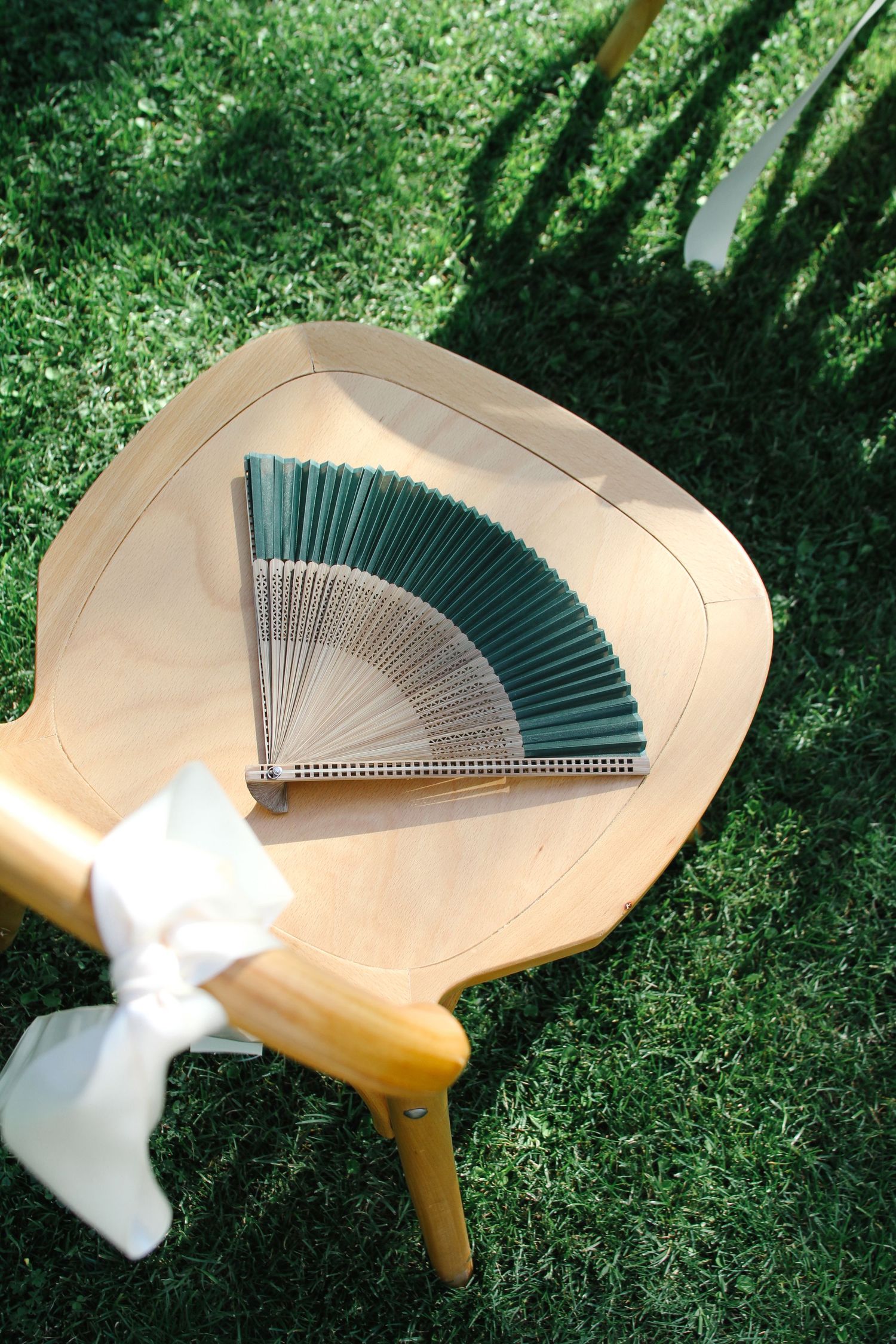 A green folding hand fan rests on a wooden chair outdoors on grass in natural sunlight.
