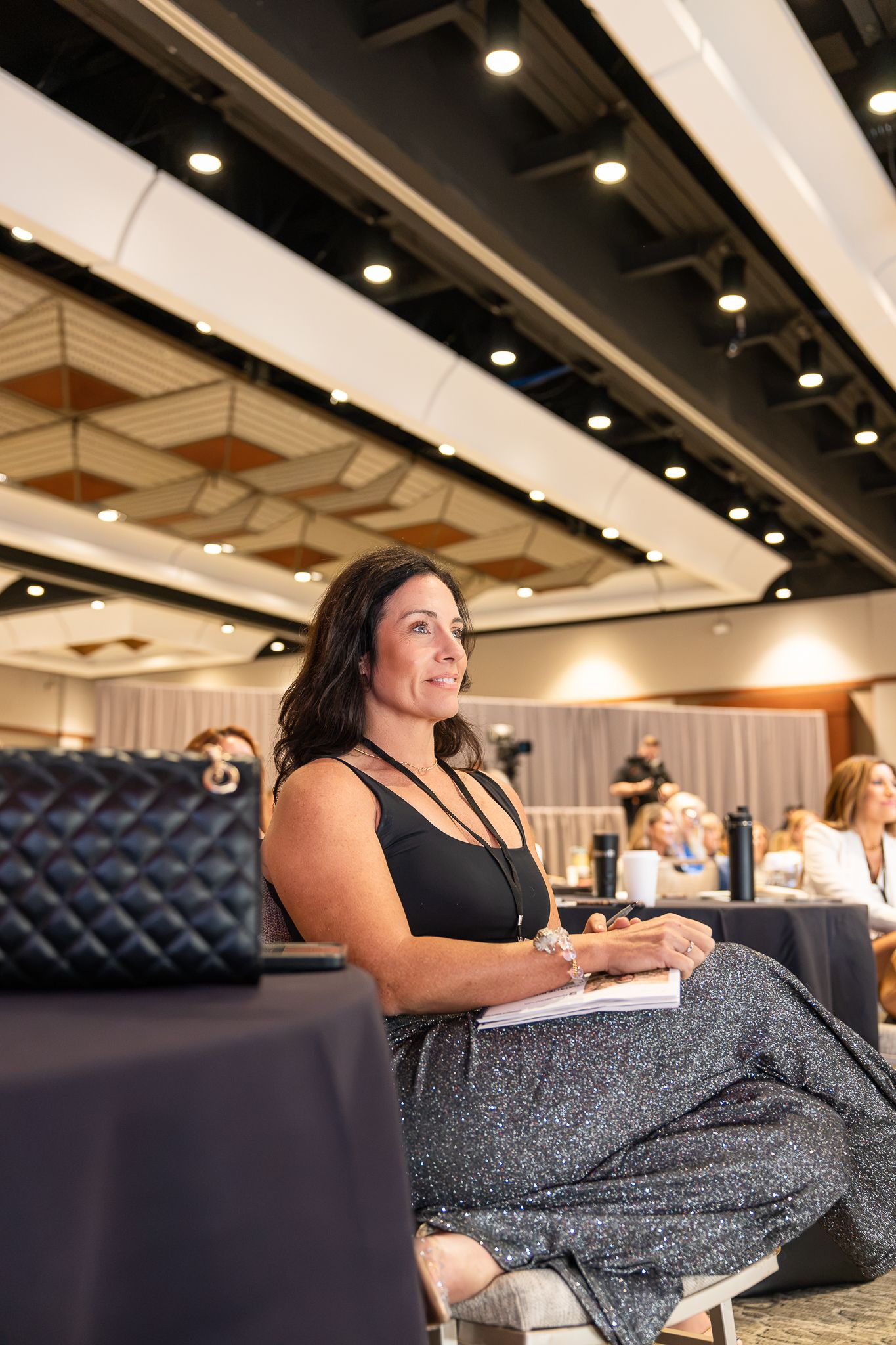 Woman in black top and sparkly skirt sits attentively at a conference, holding a notebook, with a quilted bag nearby.