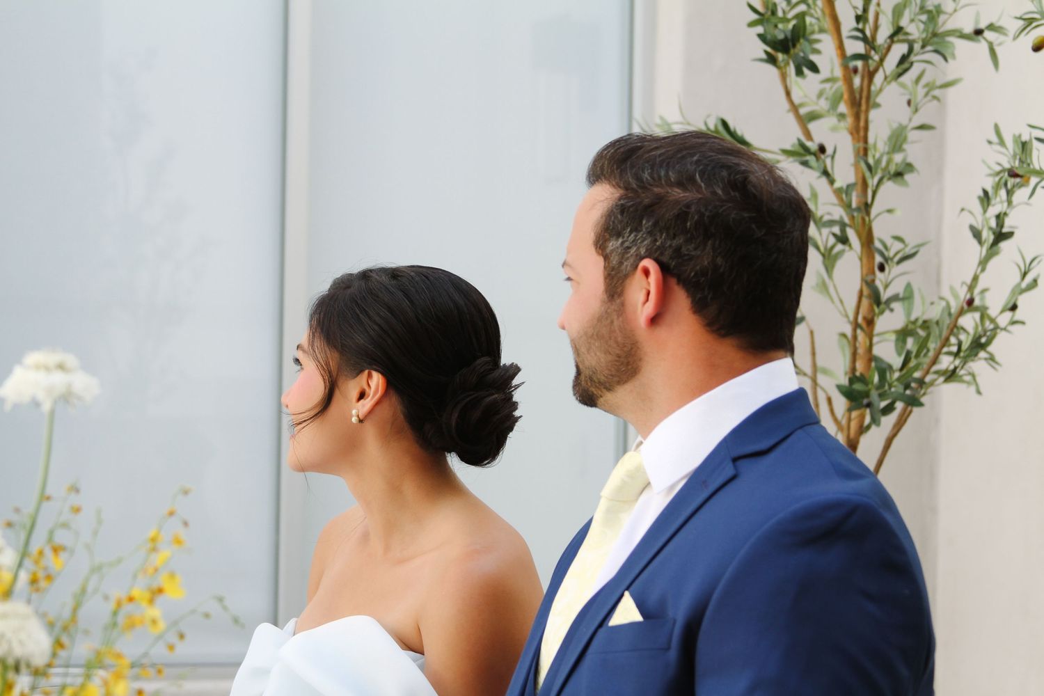 Side profile of a couple in formal wedding attire standing against greenery.