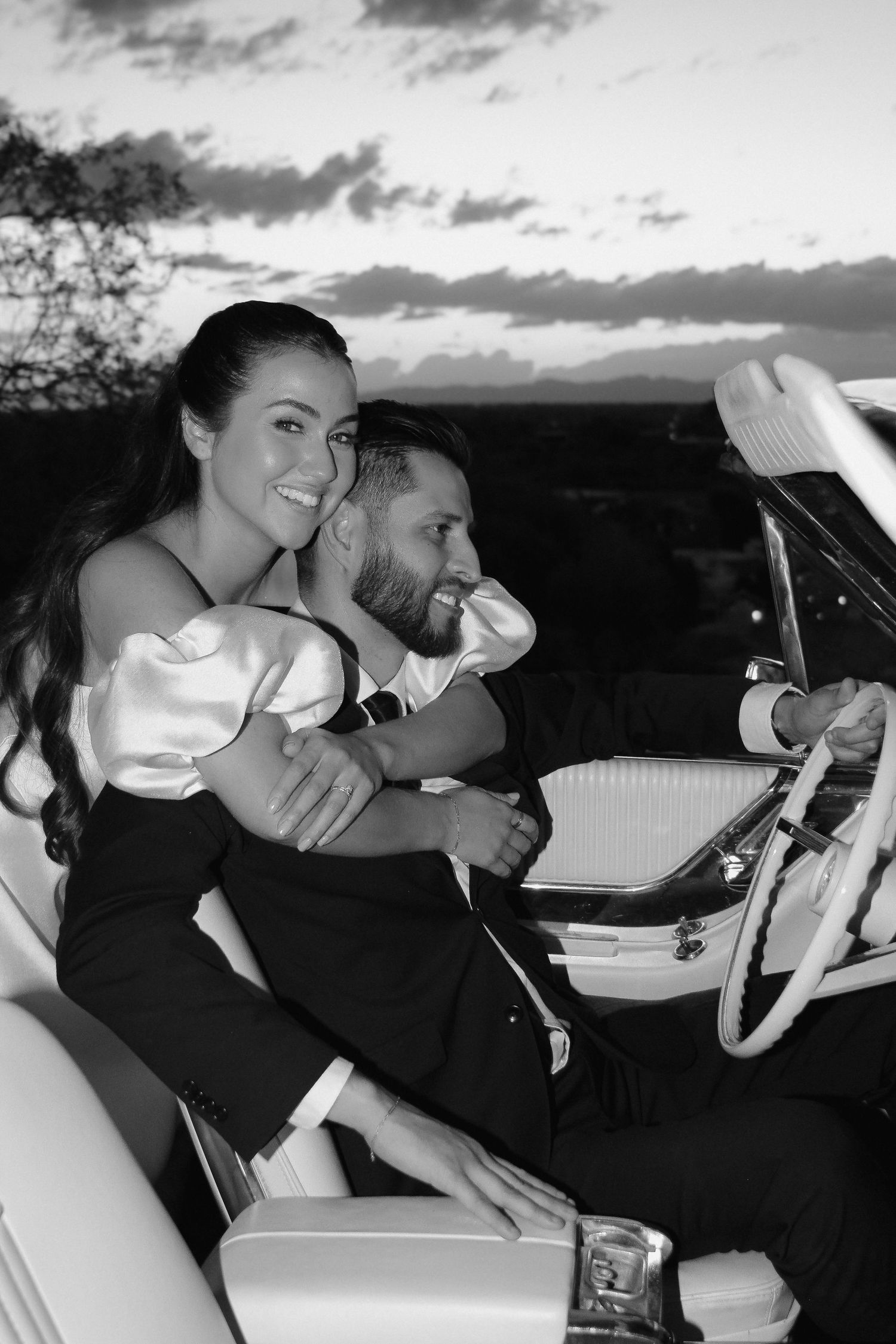 A black and white photo of newlyweds sharing an intimate moment in a classic car at sunset.