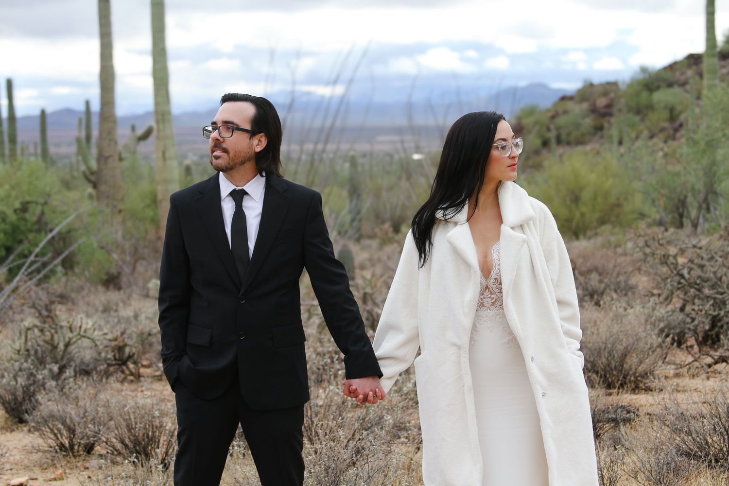 A couple holds hands while walking through a desert landscape with saguaro cacti and mountains in the background.