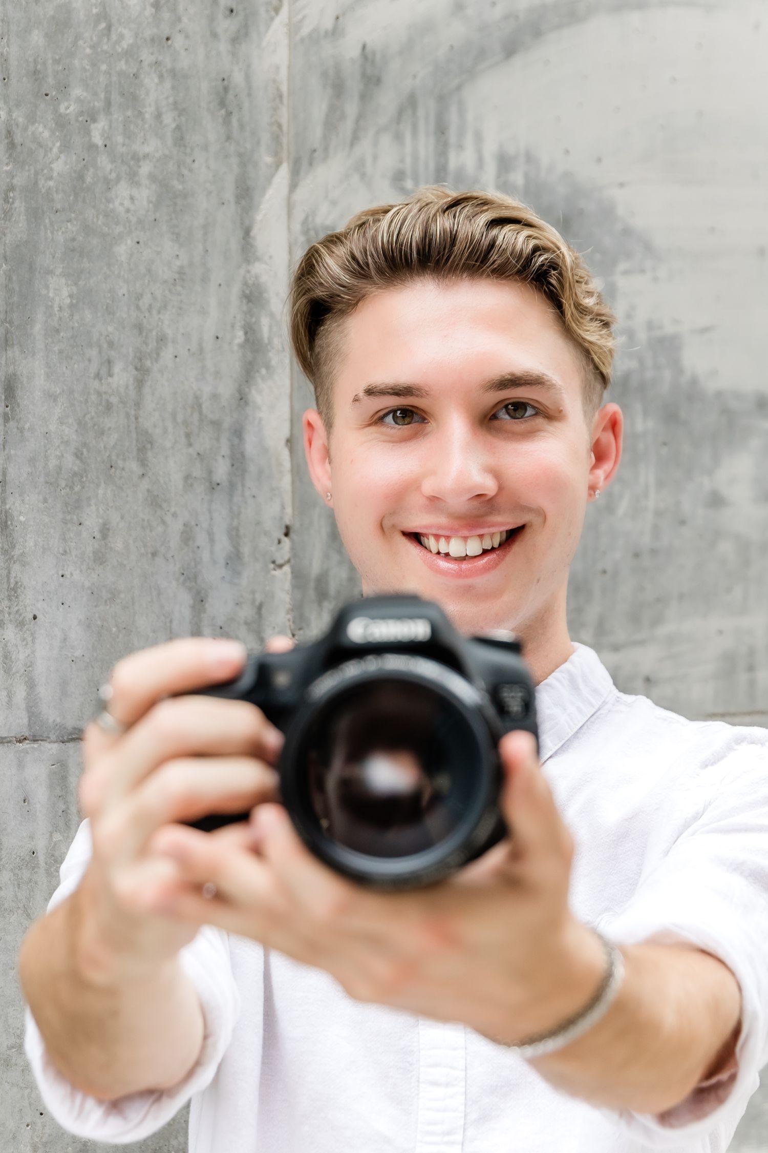 Photographer in white shirt holding camera against concrete wall.