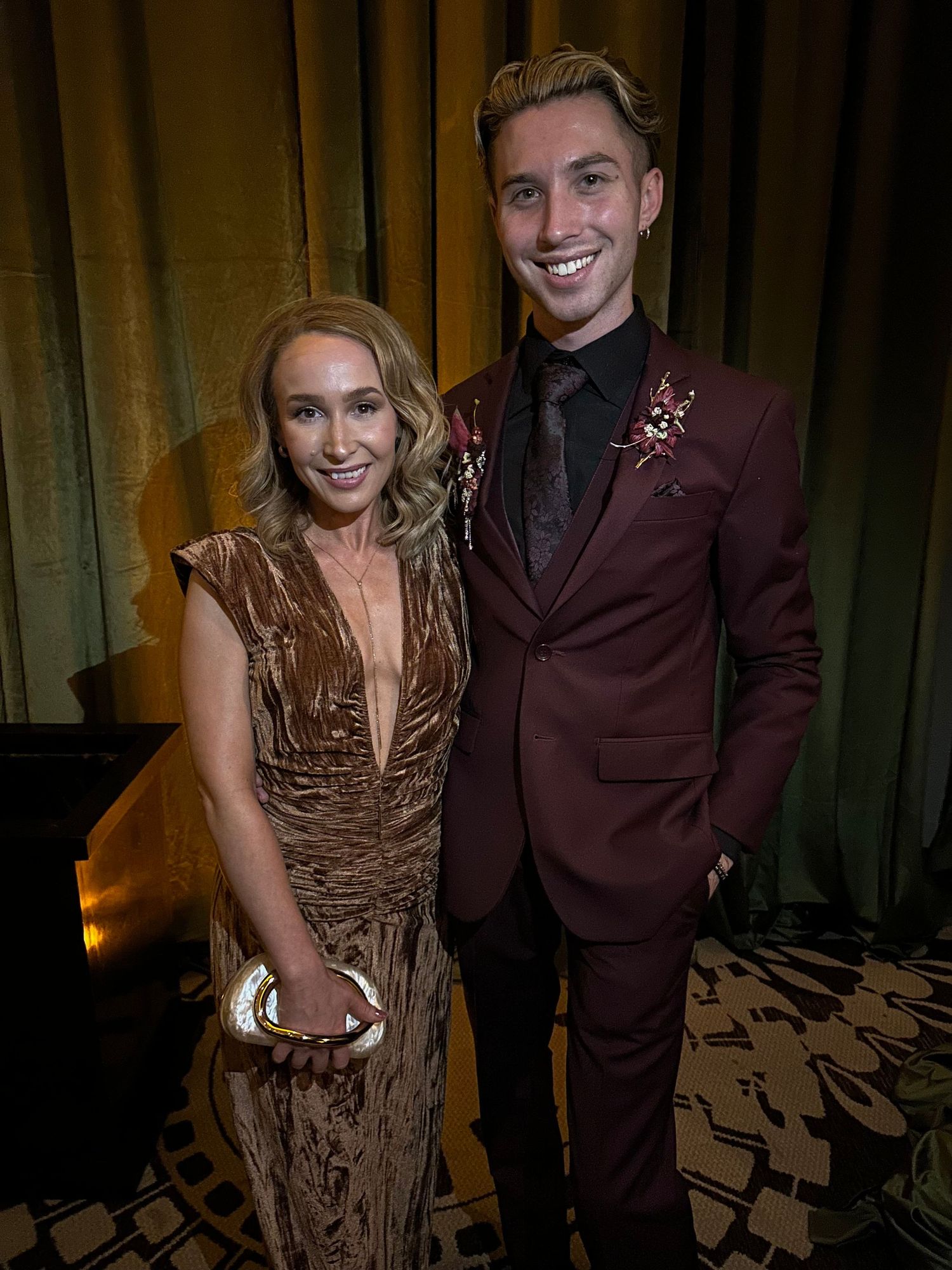 Couple posing at a formal event, woman in gold velvet gown, man in burgundy suit with floral pin.