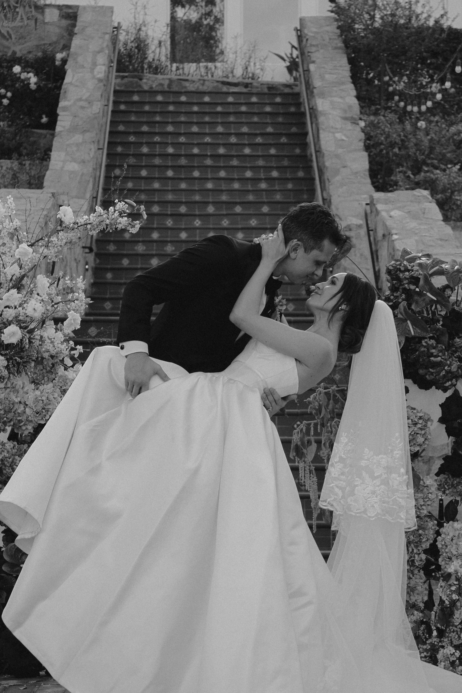 Black and white photo of a bride and groom sharing a romantic dip kiss on outdoor stairs surrounded by floral arrangements.