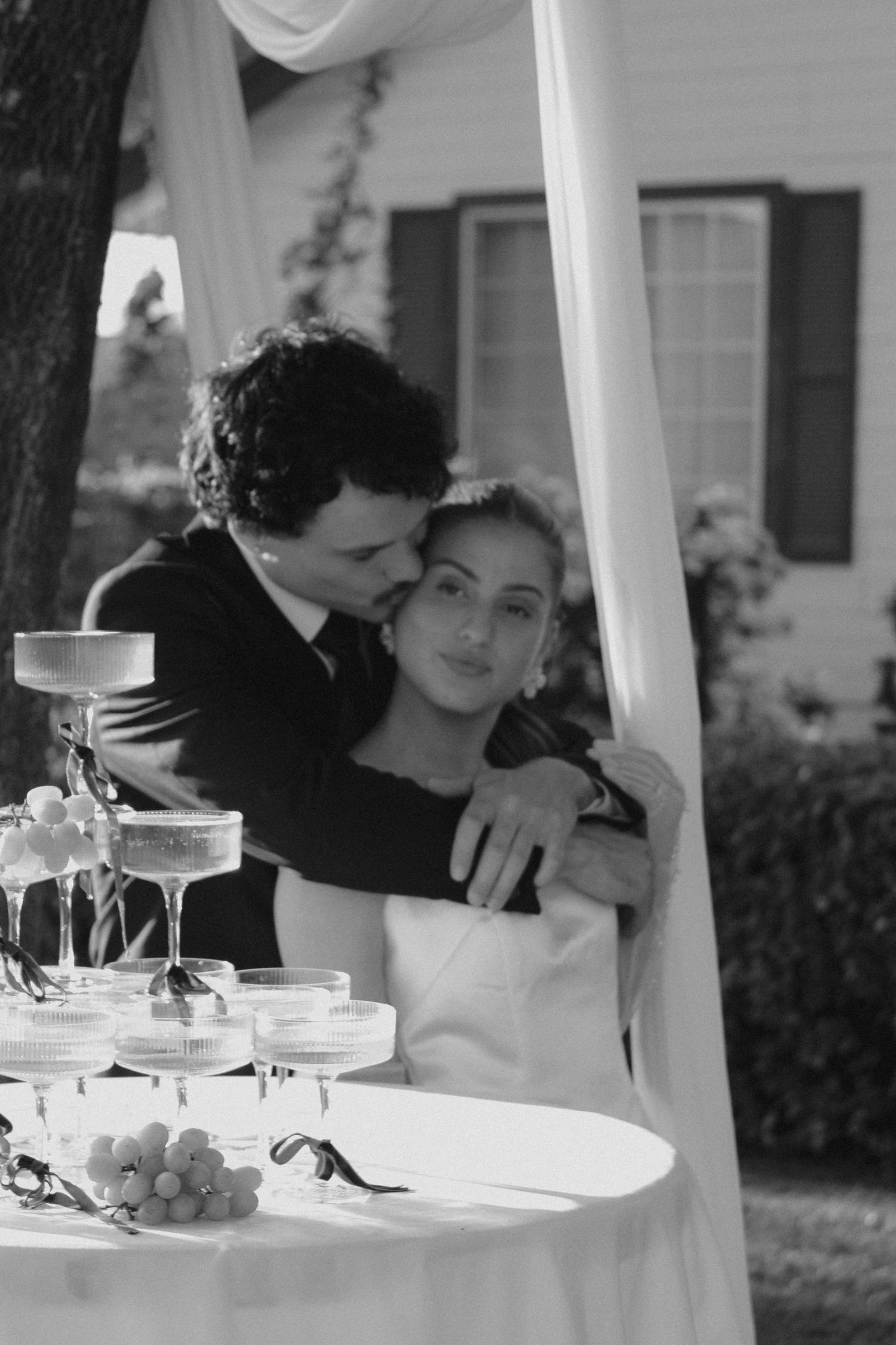 Black and white photo of a bride and groom embracing near a champagne tower at their outdoor wedding reception.