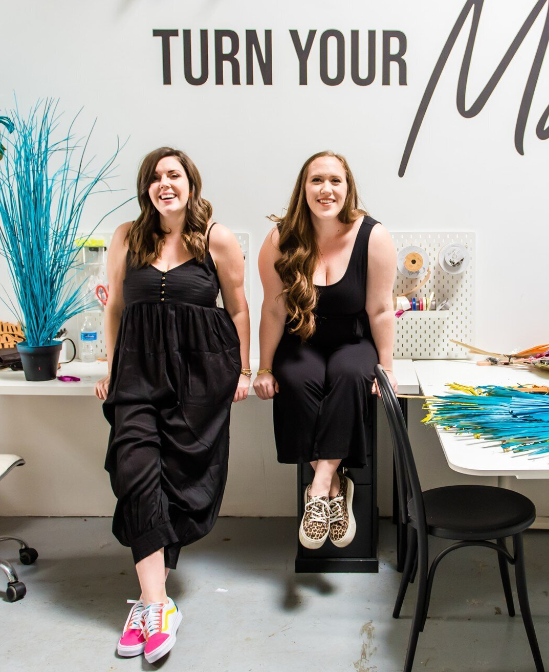 Two women in black dresses sitting casually in modern office space with motivational wall decor.