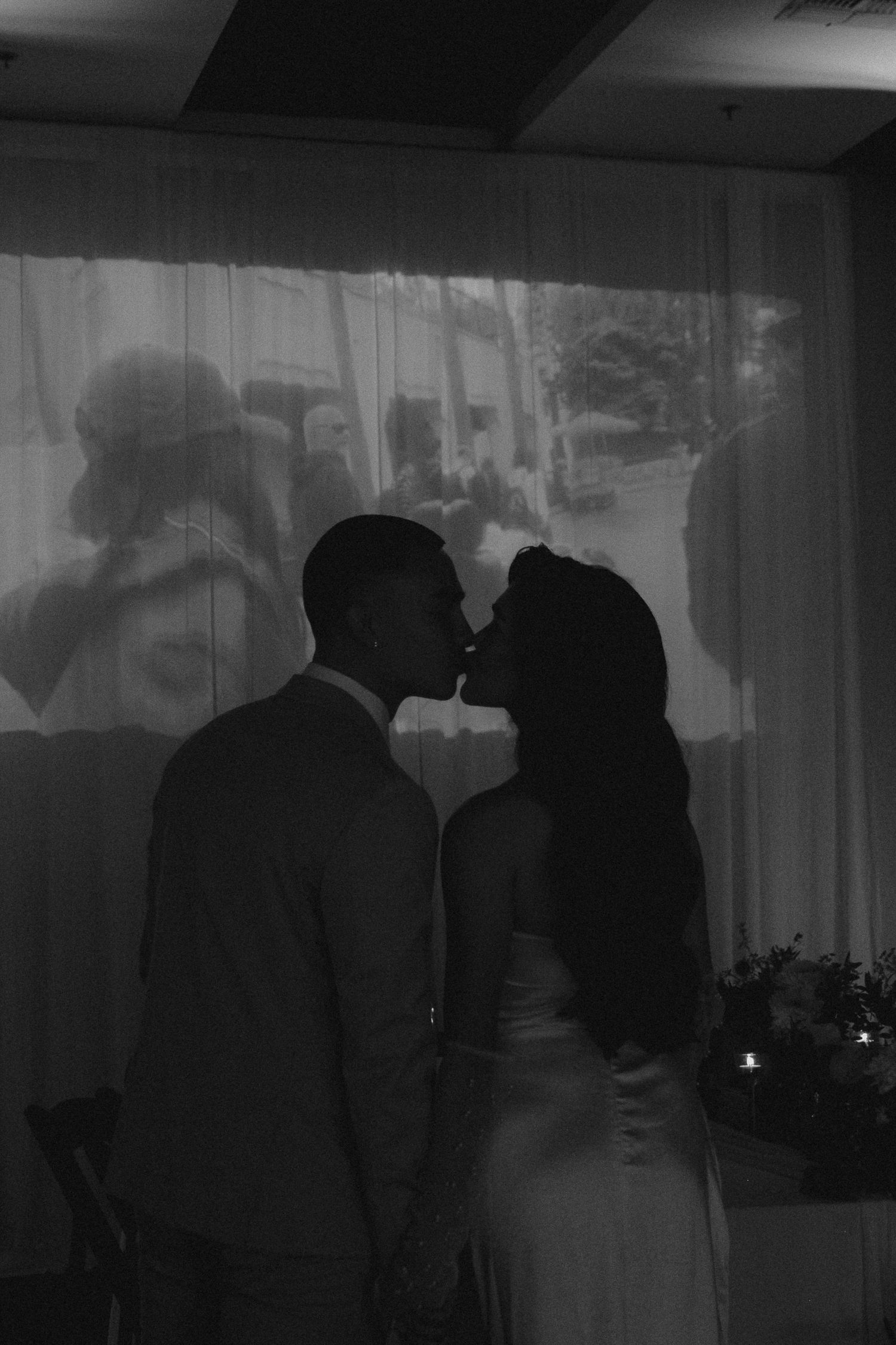 Black and white photo of a couple sharing a romantic kiss at their wedding reception with a projected image behind them.