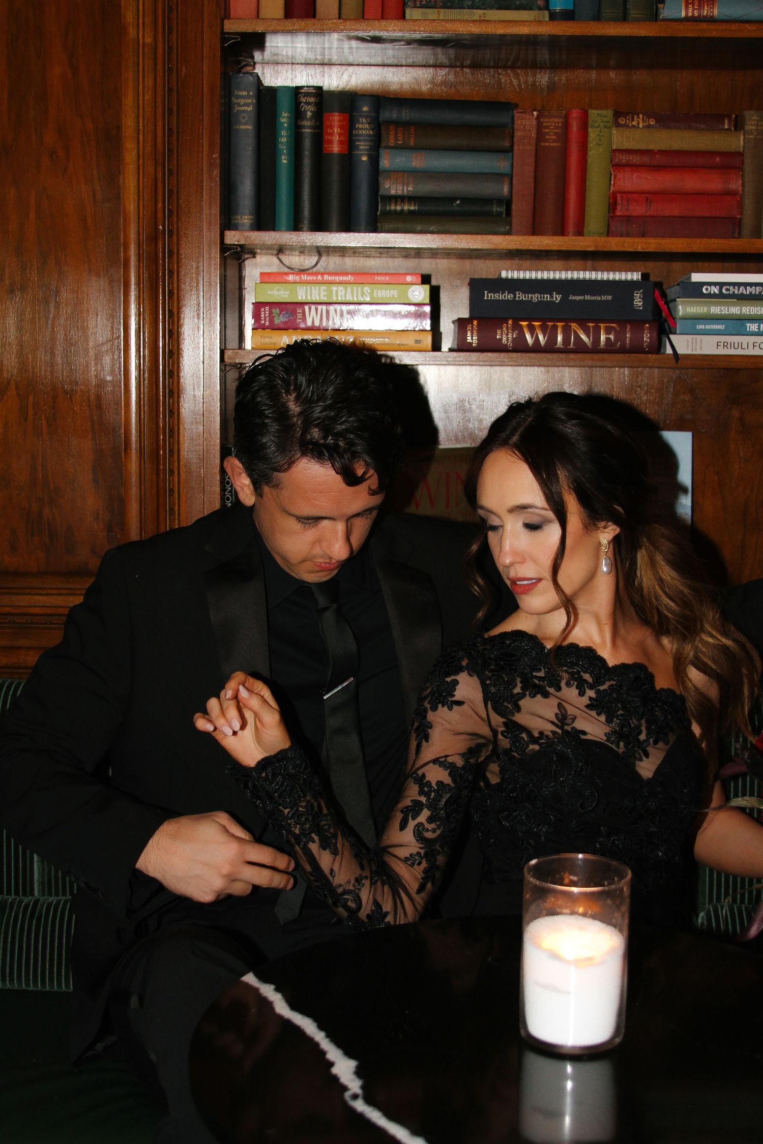 A couple in elegant black attire sharing an intimate moment at a table with a candle and bookshelf backdrop.