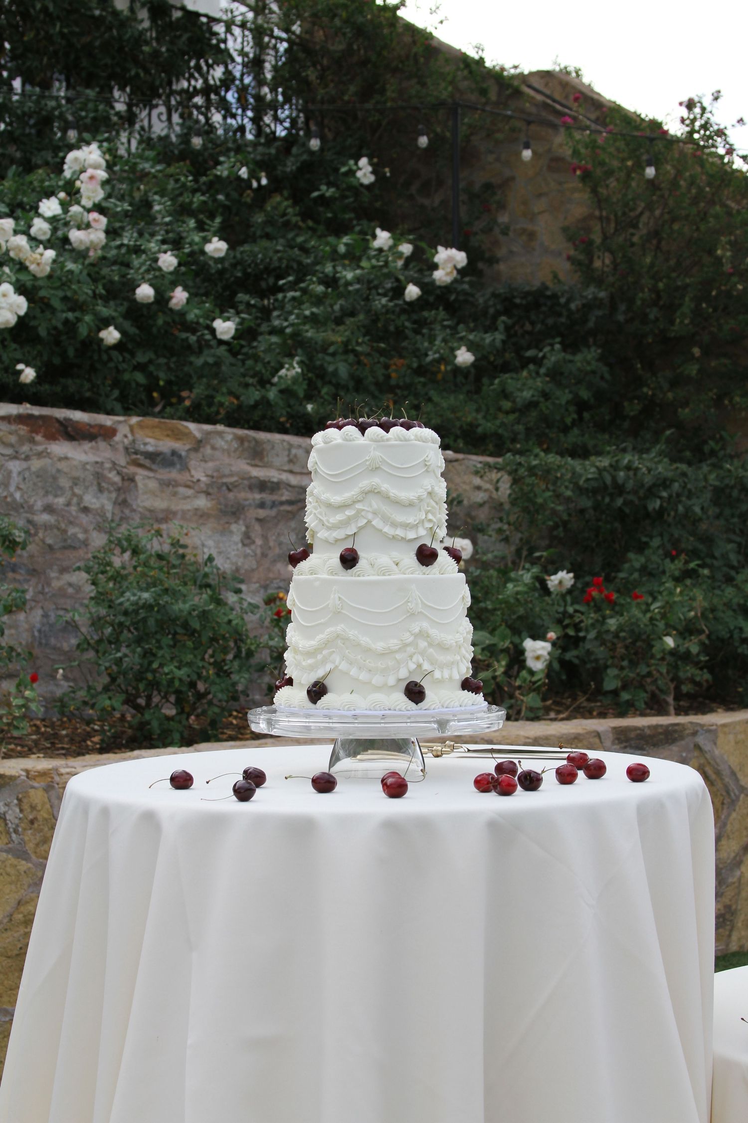 Three-tiered white wedding cake decorated with red cherries displayed on a white linen table.