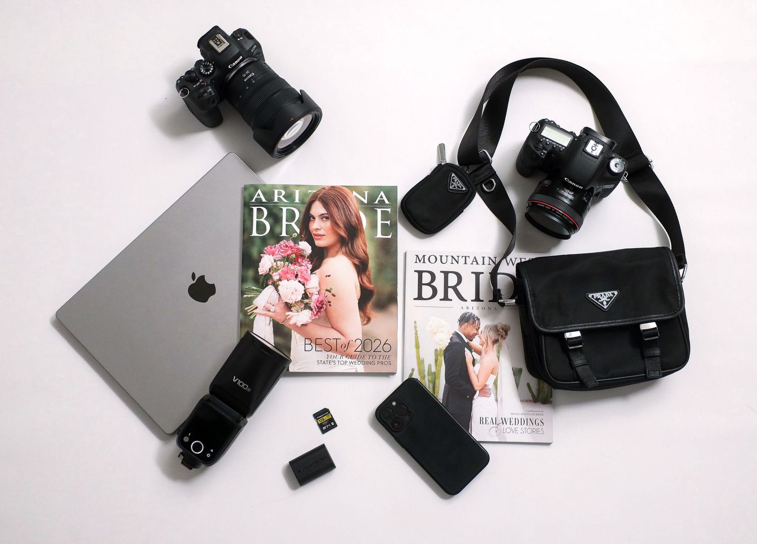 Flat lay of two DSLR cameras, MacBook, Arizona Bride magazines, Prada bag, smartphone, and memory cards on white background.