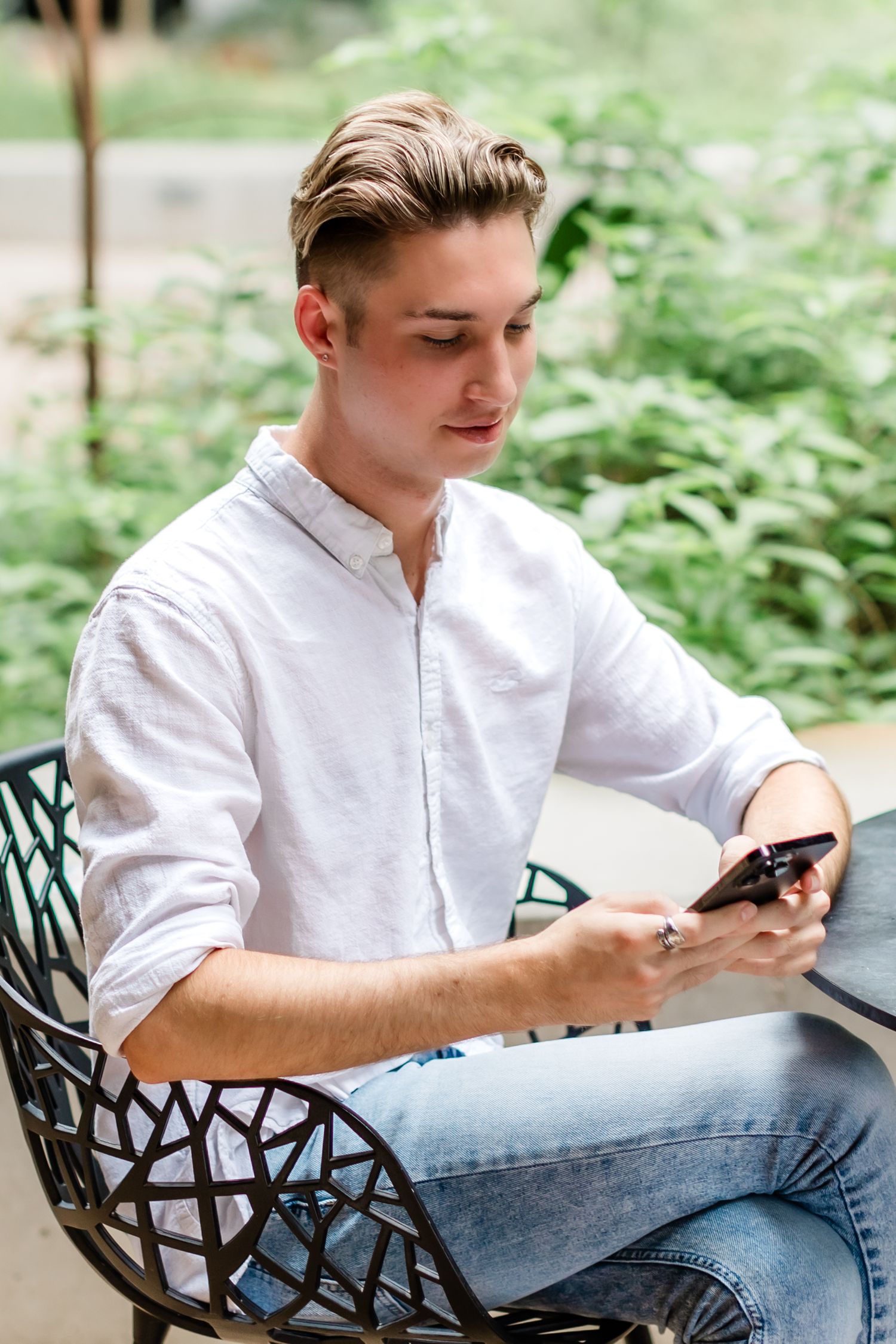 Young adult in white button down shirt looks at phone while sitting outdoors.