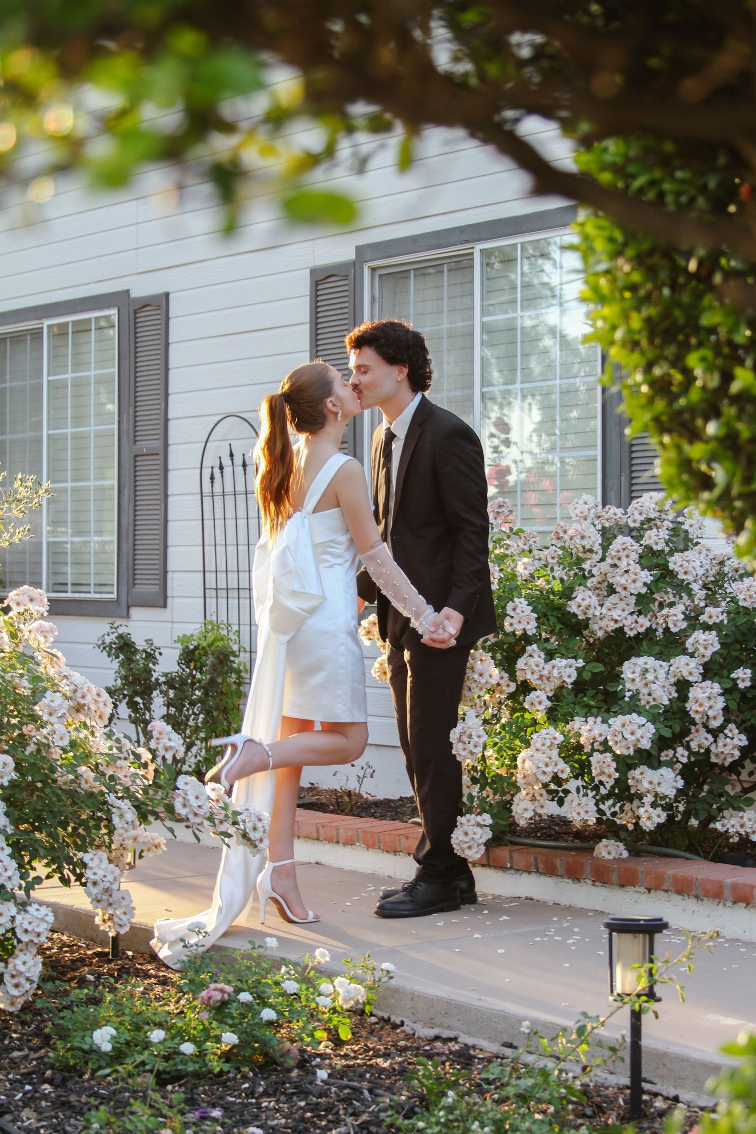 Couple sharing a romantic kiss in a white dress and dark suit among blooming white roses outside a charming home.