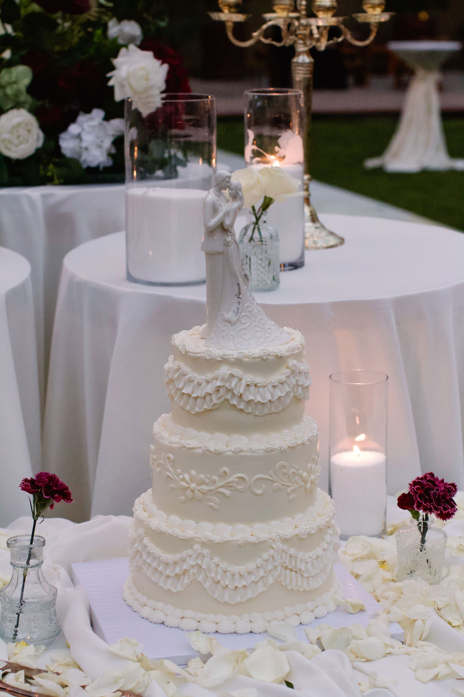 Elegant white wedding cake with lace piping details and pearl accents displayed on table with candles and floral decorations.