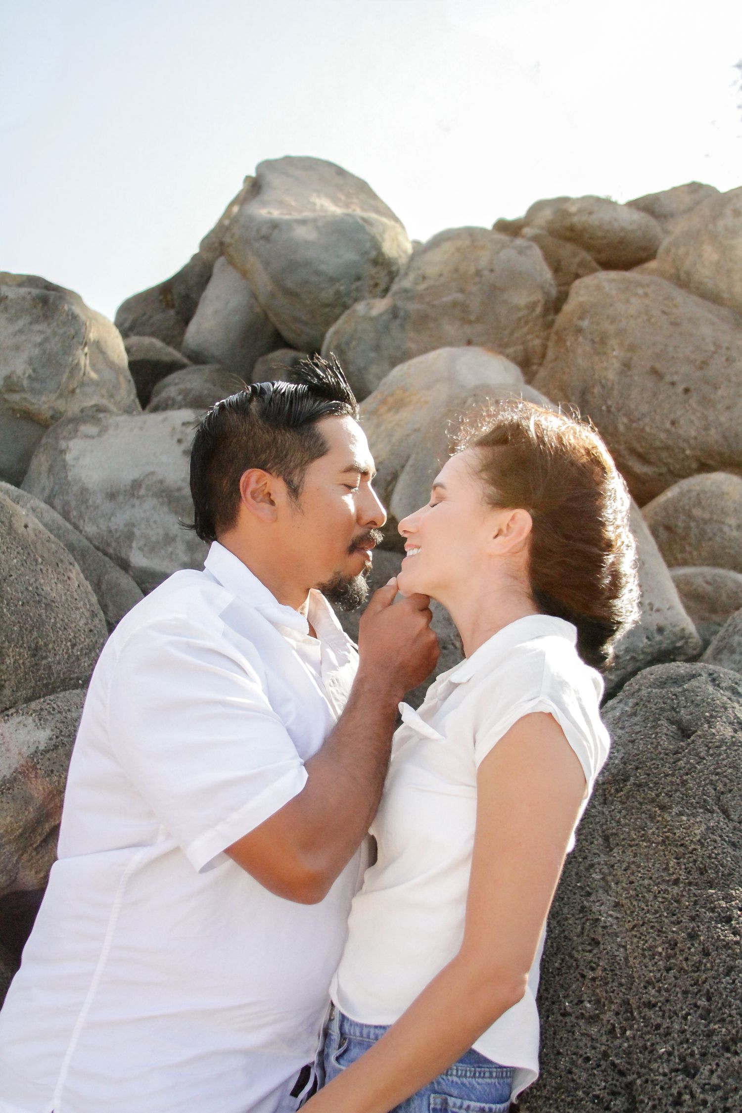 Romantic couple sharing an intimate moment against large granite boulders in sunlit outdoor setting.