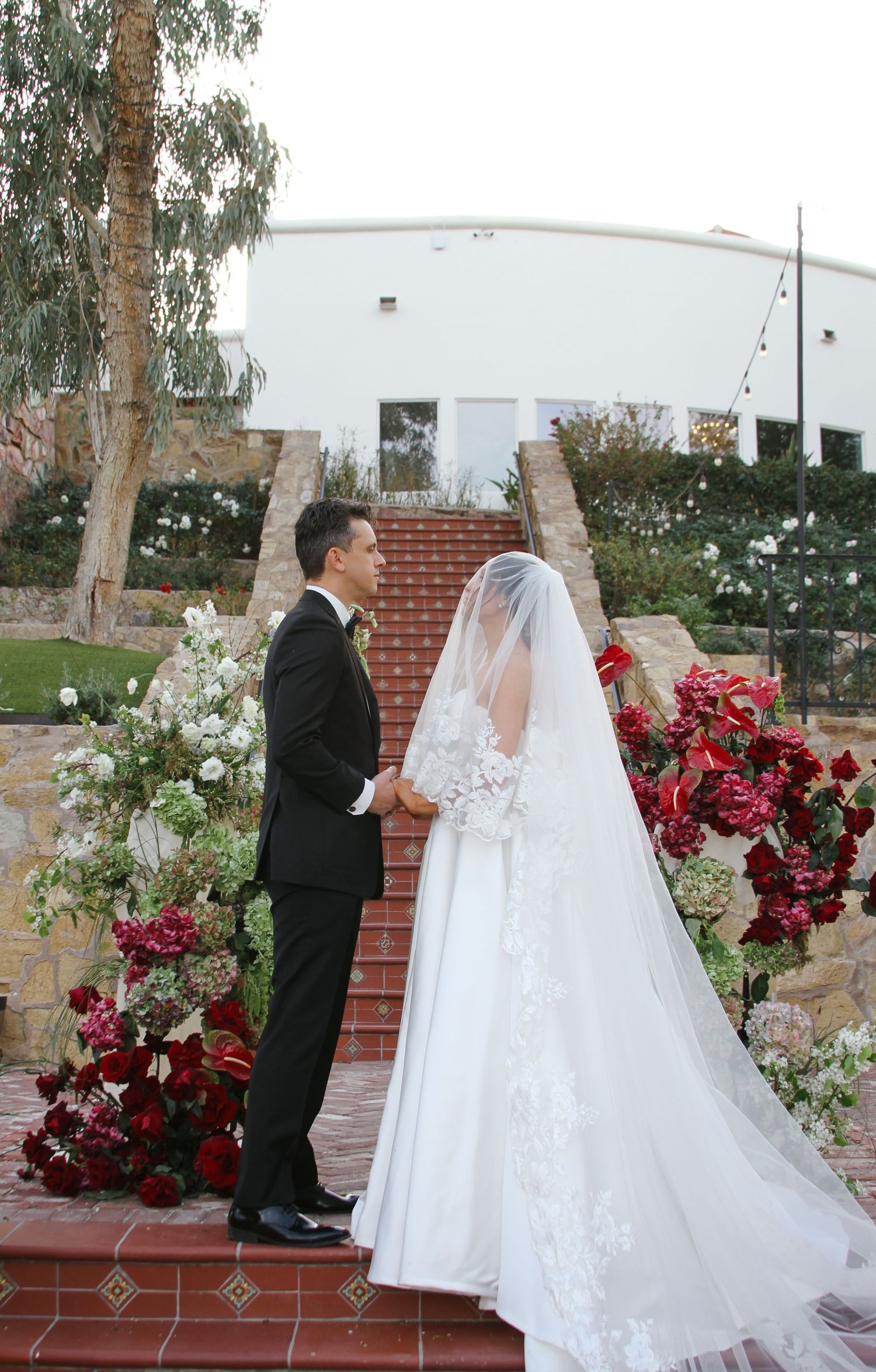 Bride and groom exchange vows outdoors surrounded by lush red and white floral arrangements at an elegant wedding ceremony.