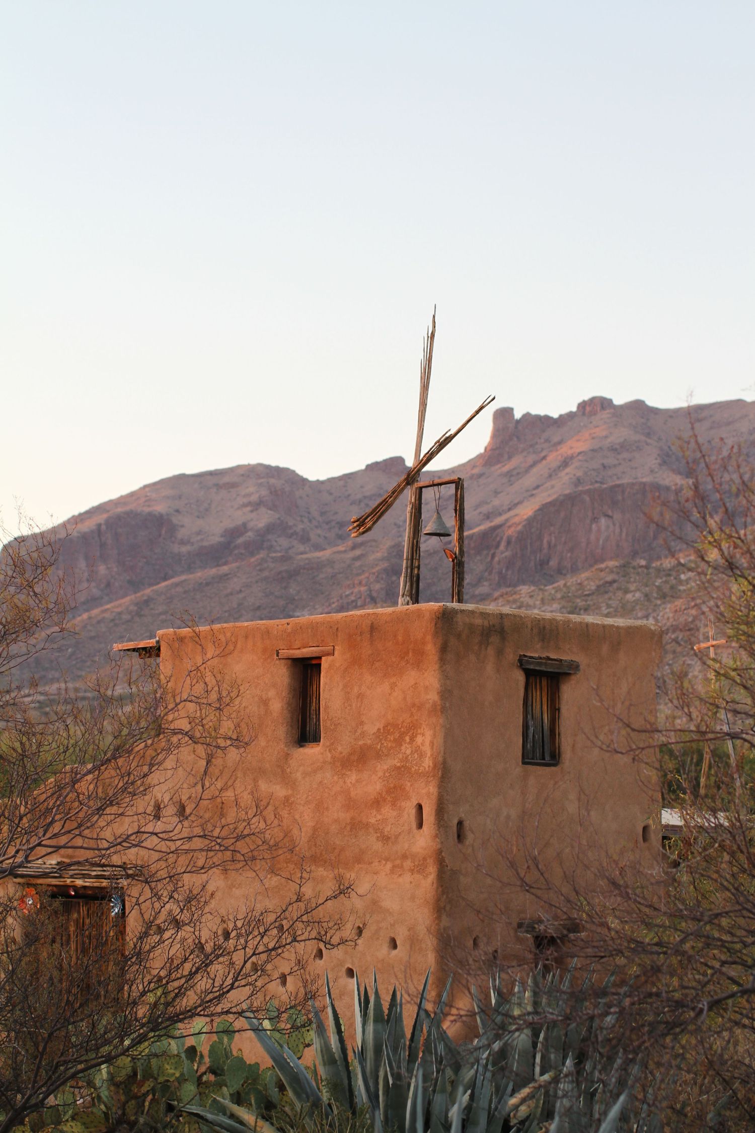 Adobe chapel with cross on roof stands against desert mountain backdrop at sunset.